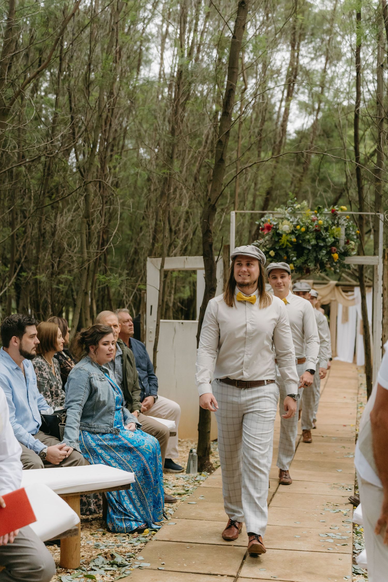 Wedding ceremony procession on a wooden walkway in a forest setting with seated guests and floral arch decor.