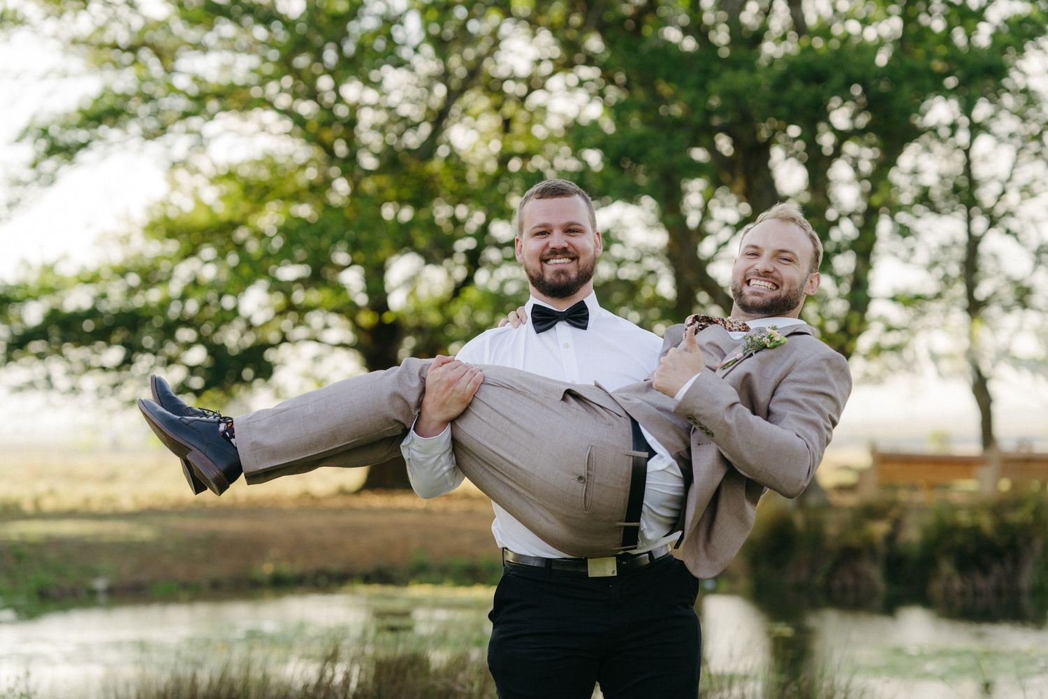 Wedding couple playfully poses with groom carrying guest against scenic outdoor backdrop.