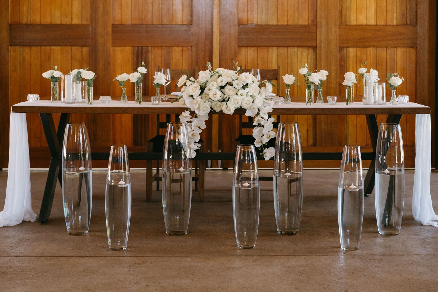 Modern wedding reception table with clear ghost chairs and white floral centerpiece against wooden paneled wall.