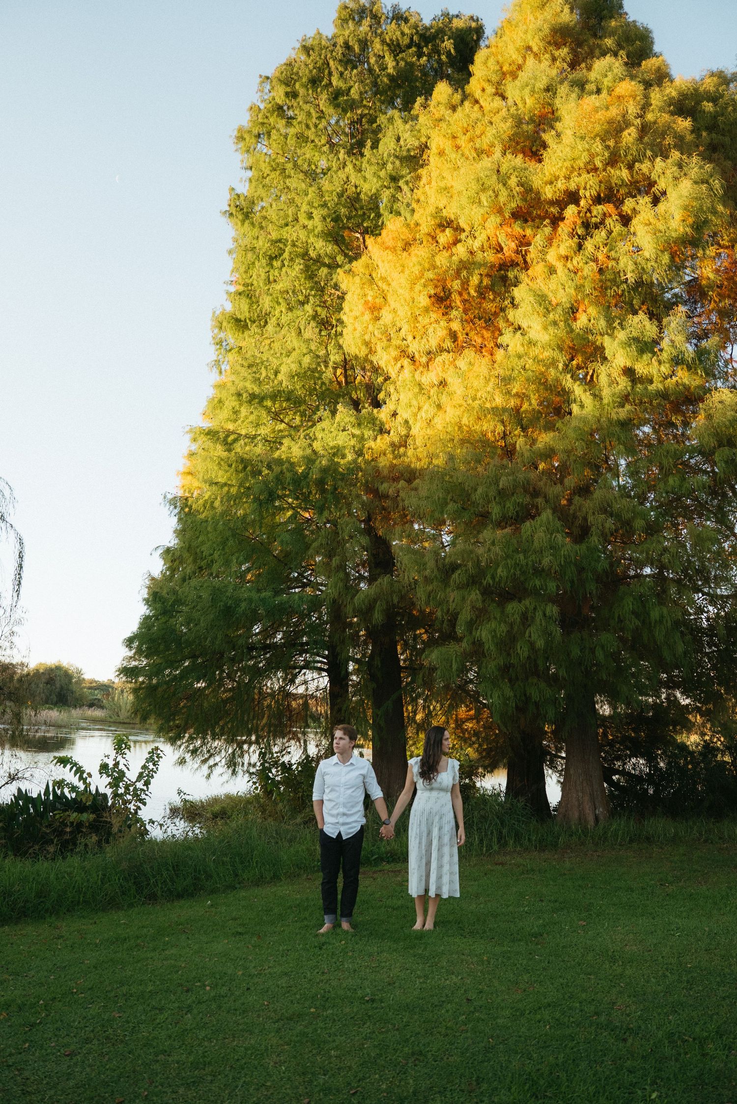 A couple stands together on a grassy lawn at sunset with golden autumn trees and a lake in the background.