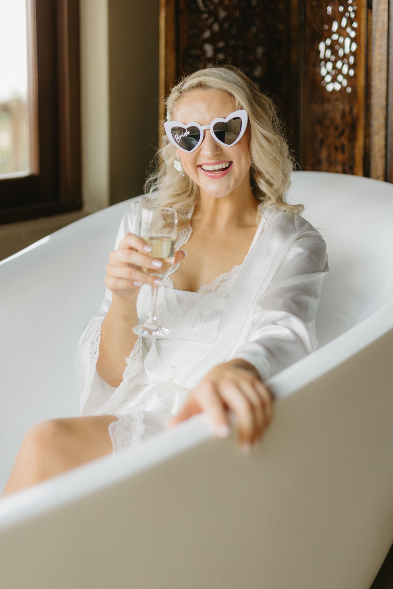 Person relaxing in a white bathtub wearing heart-shaped sunglasses and holding a champagne glass.