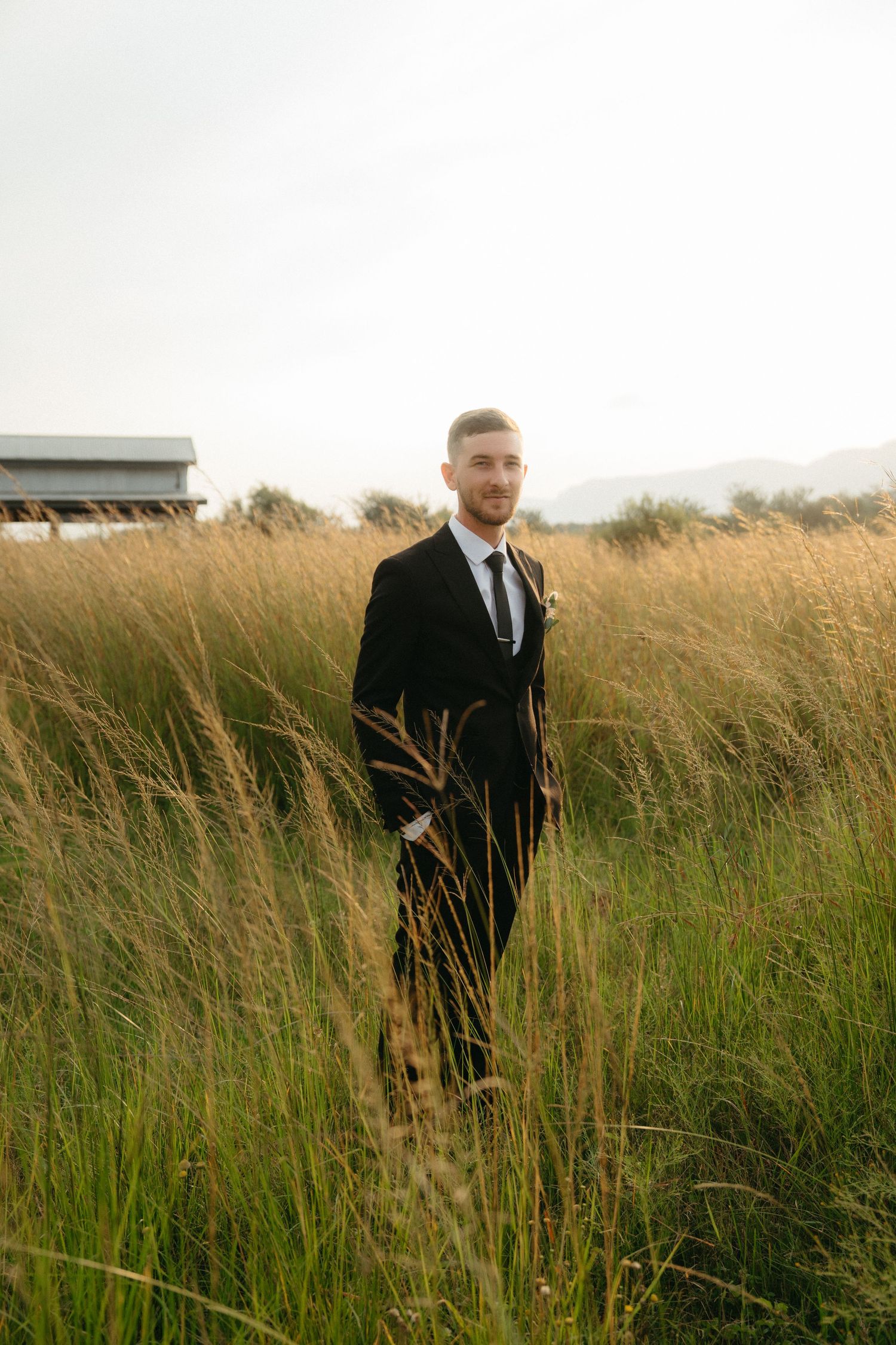 A groom in a black suit stands in a field of tall golden grass at sunset.