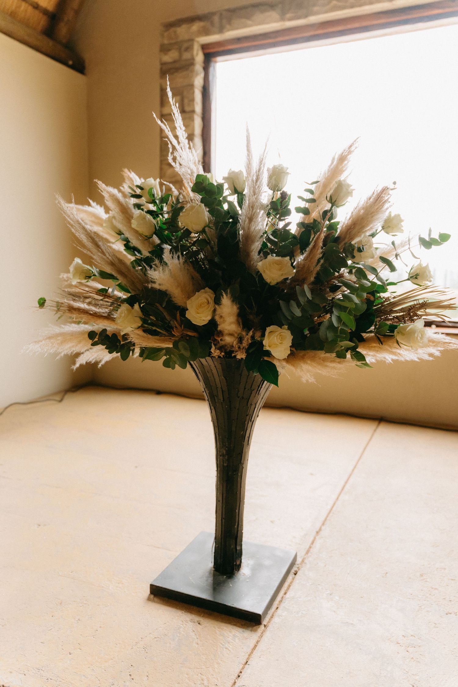 Elegant floral arrangement with pampas grass and greenery in a tall vase against white backdrop.