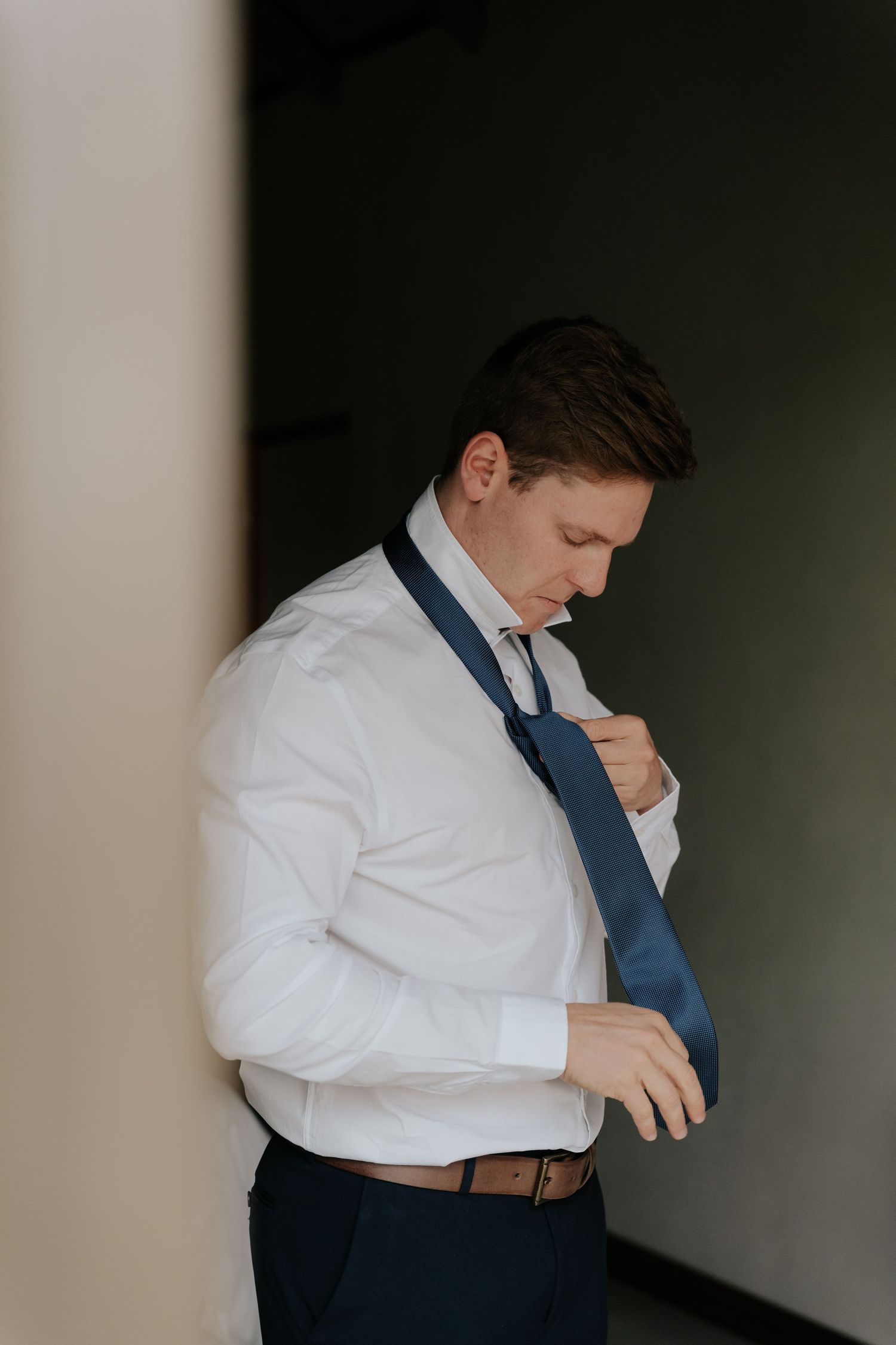 Groom adjusting his navy blue tie while getting ready in a dimly lit room.