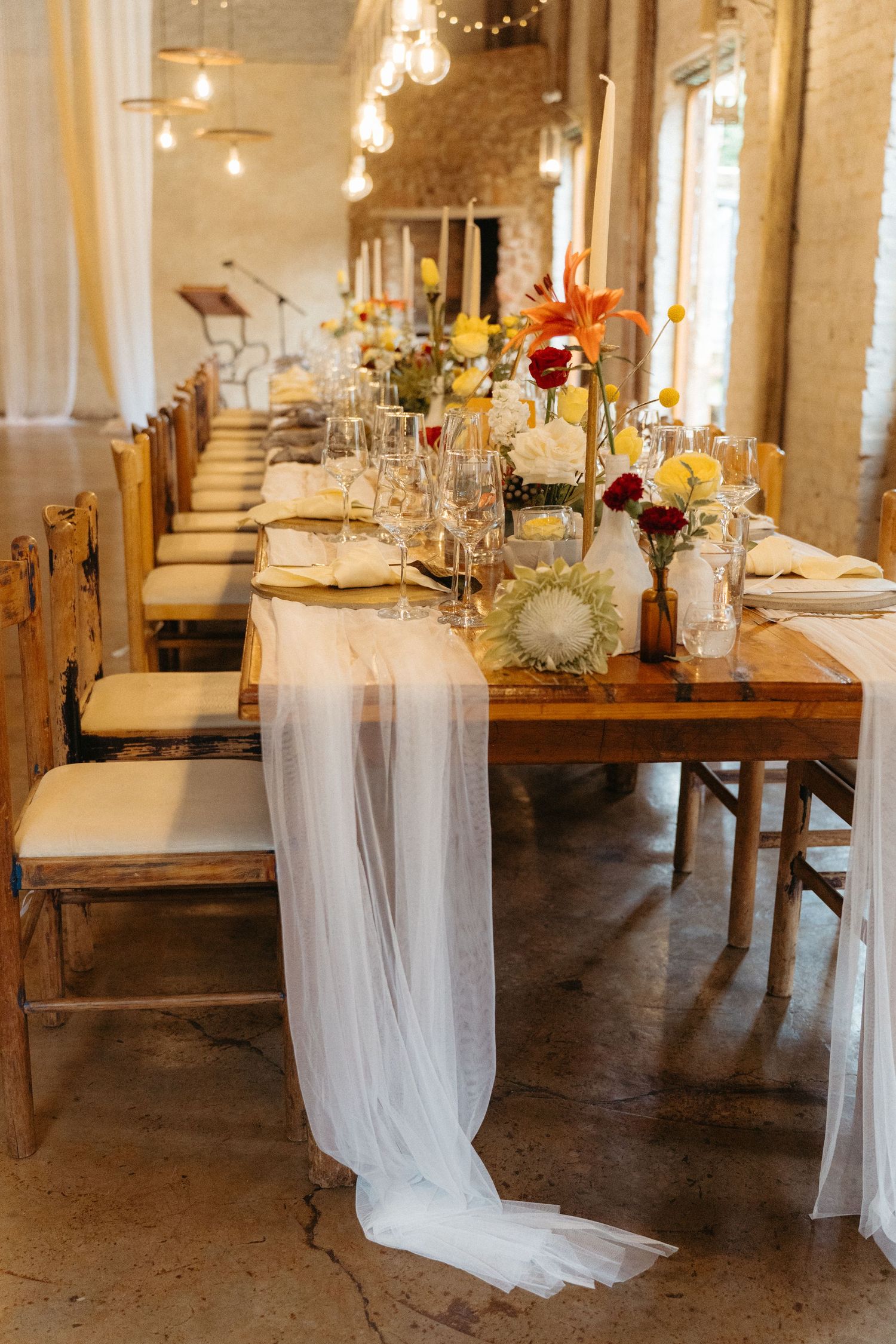 Elegant rustic wedding reception table with floral centerpieces, flowing white fabric, and string lights in a brick venue.