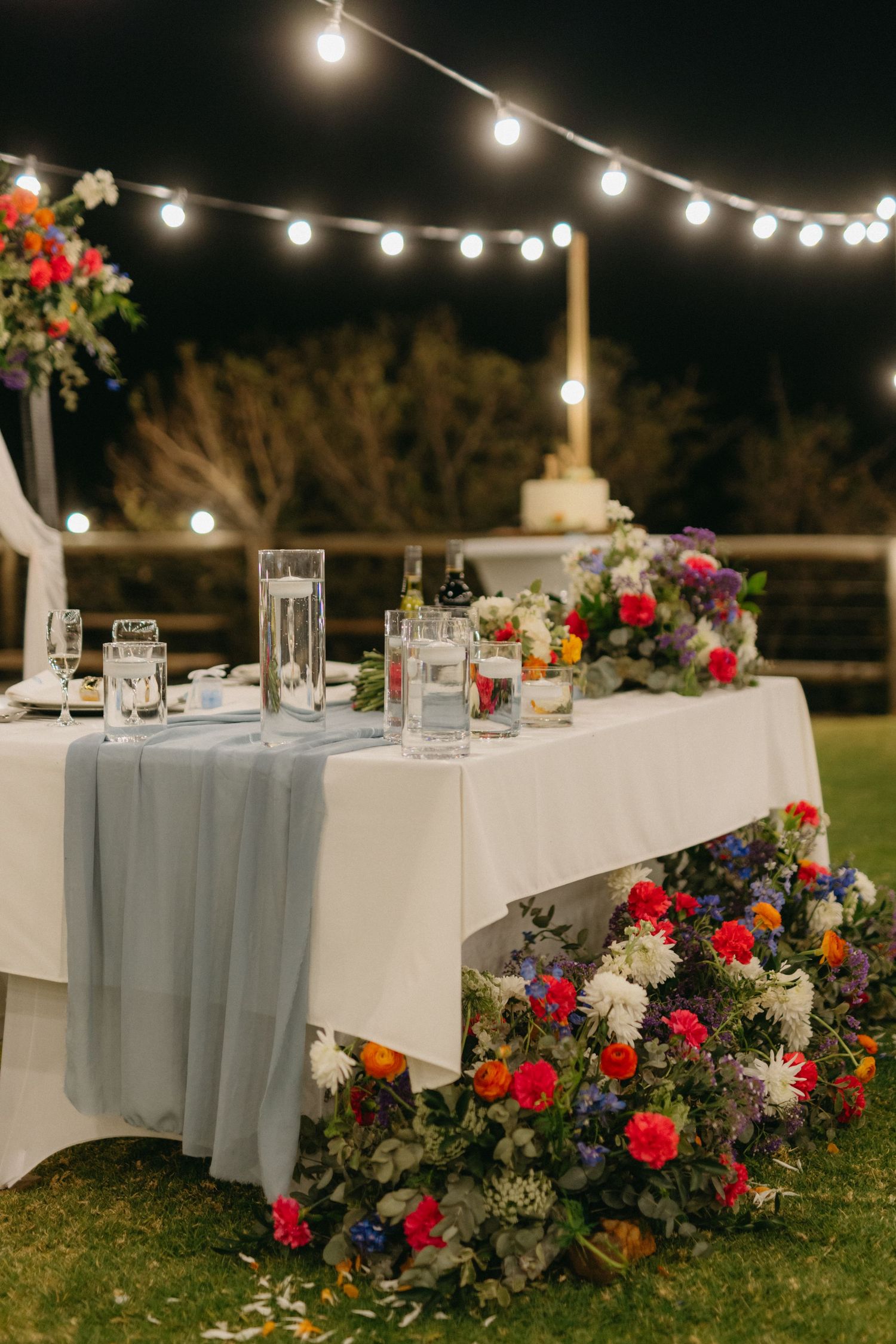 Outdoor wedding reception tables with string lights and floral arrangements at dusk.
