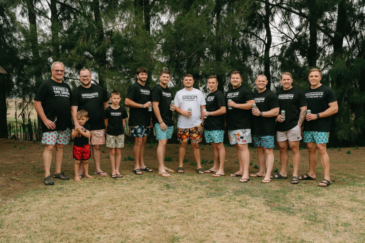 Groomsmen in matching black shirts and colorful swim trunks pose together outdoors.