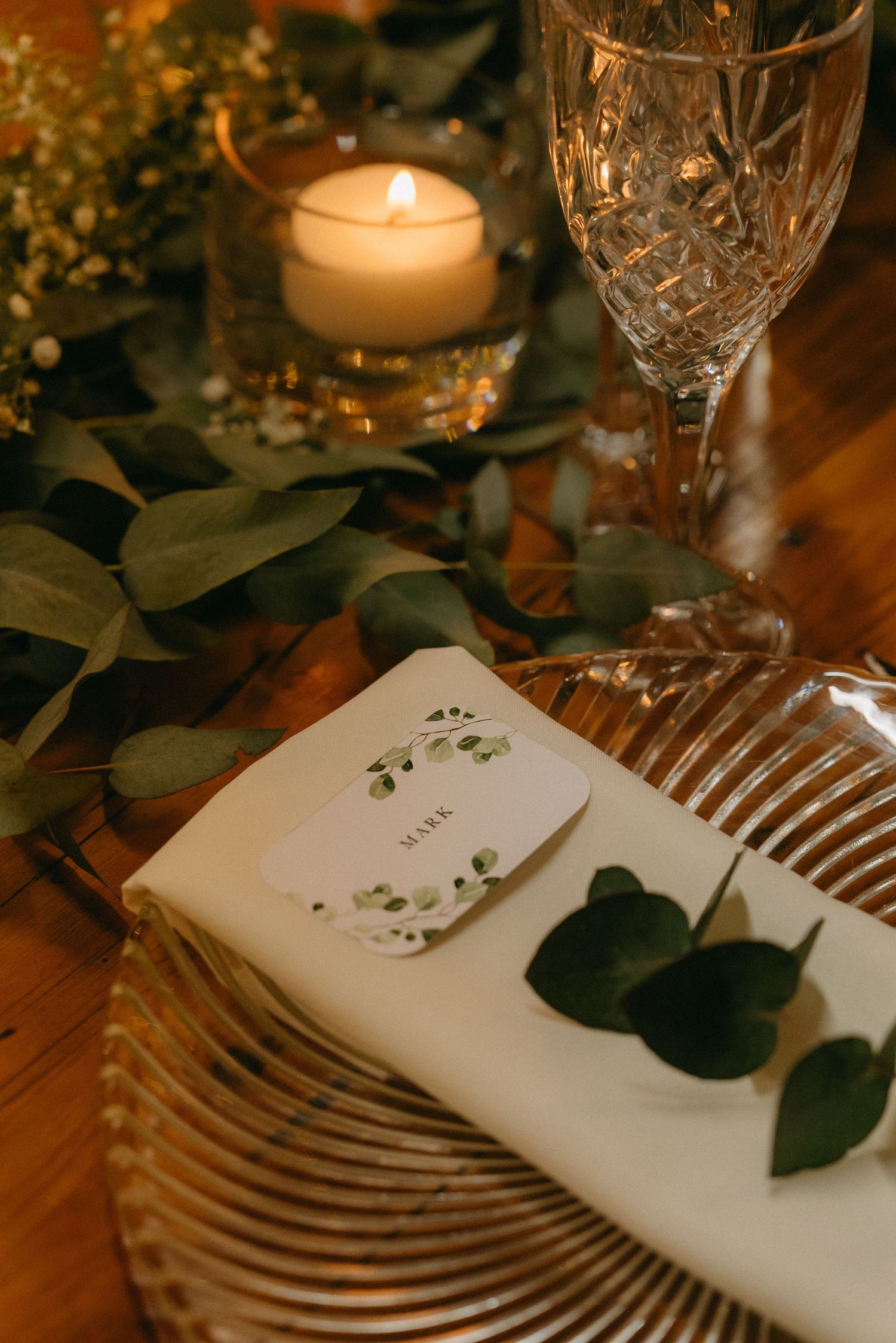 Elegant table setting with crystal glasses, white napkins, and eucalyptus leaves illuminated by candlelight.
