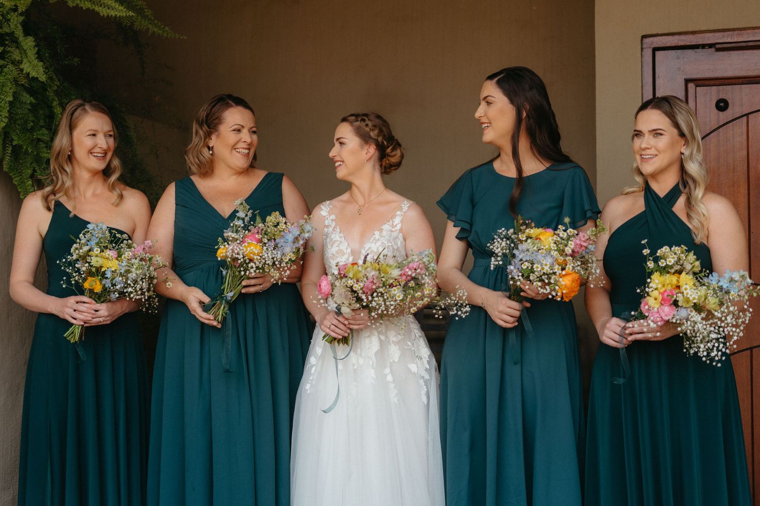 Bridal party in emerald green dresses holding colorful bouquets pose together outdoors.