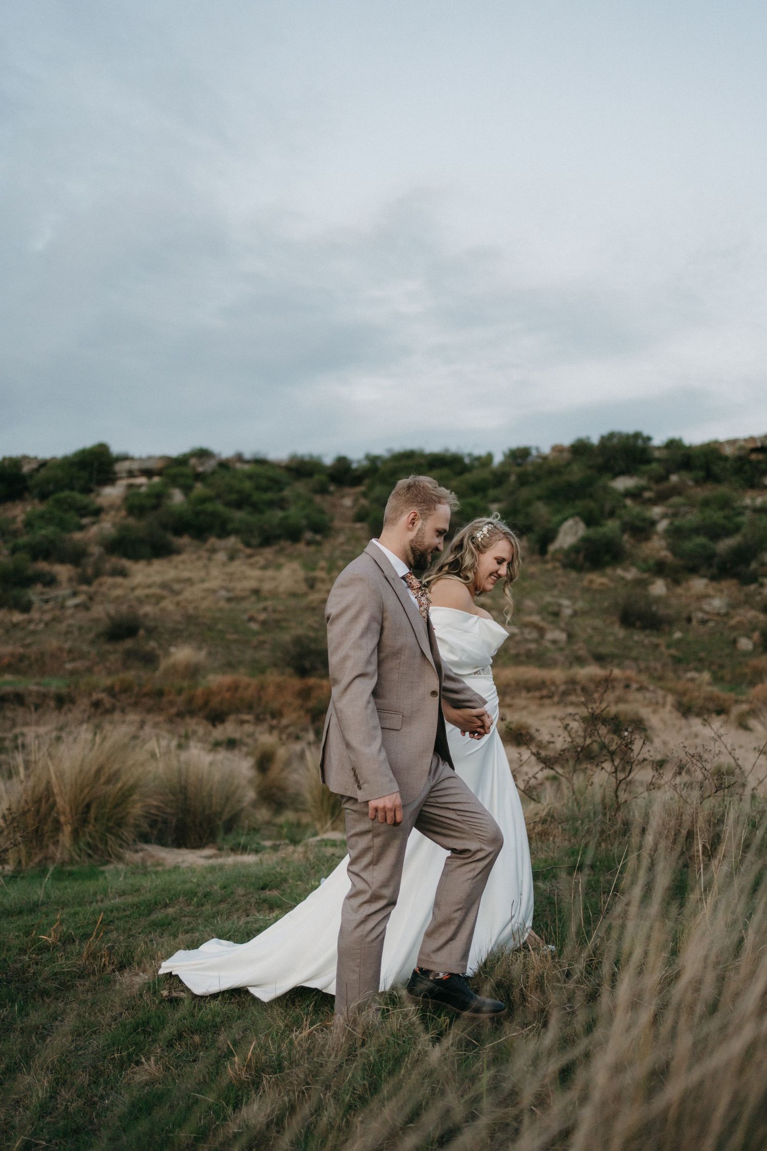 Romantic wedding couple embraces on hillside with flowing white dress and beige suit under moody sky.