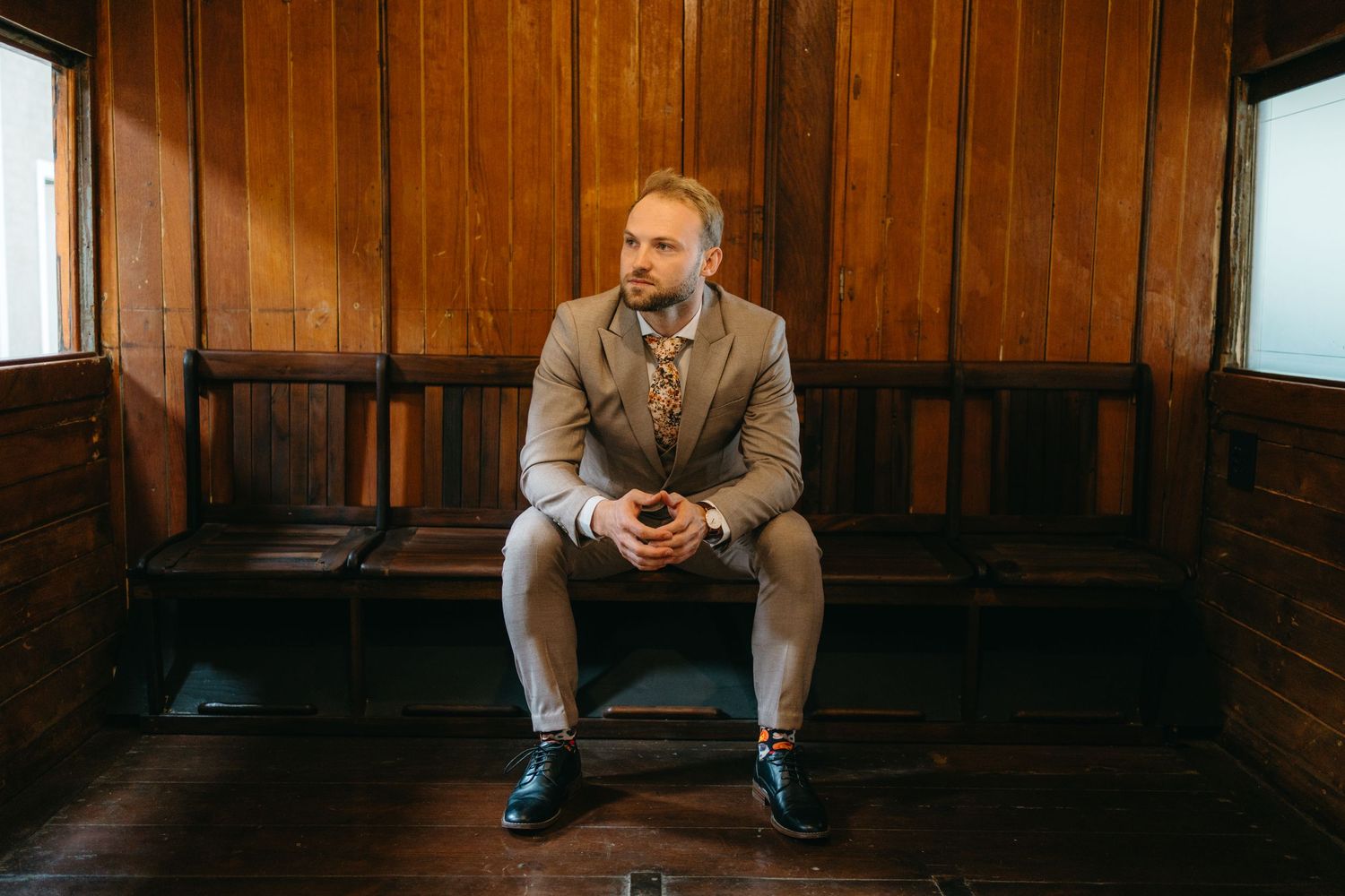 A person wearing a grey suit sits pensively on a wooden bench in a room with wooden paneled walls.
