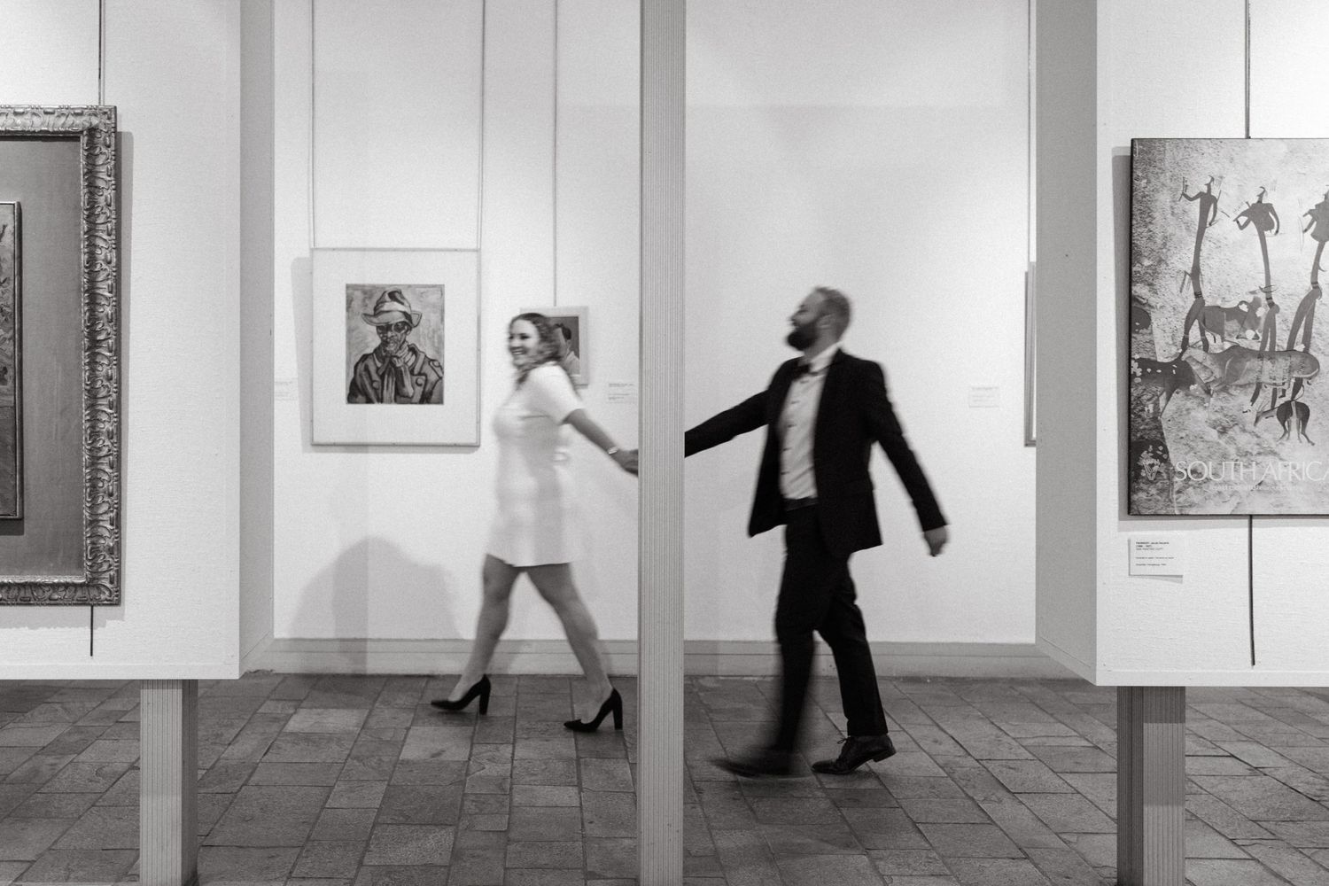 A couple dances playfully in an art gallery with framed artwork on the walls.