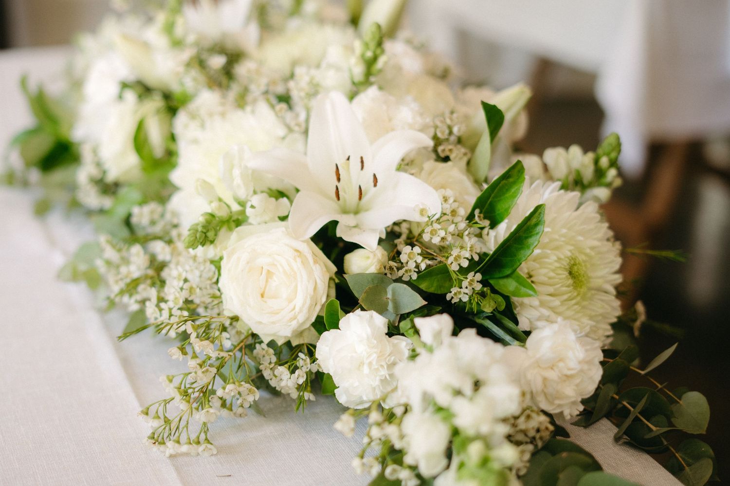 Fresh white floral arrangement featuring roses, chrysanthemums and delicate greenery on a light surface.