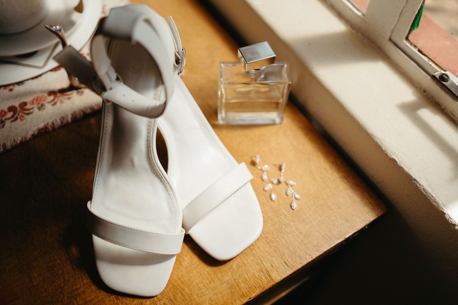 White bridal sandals and perfume bottle arranged on wooden surface in natural light.