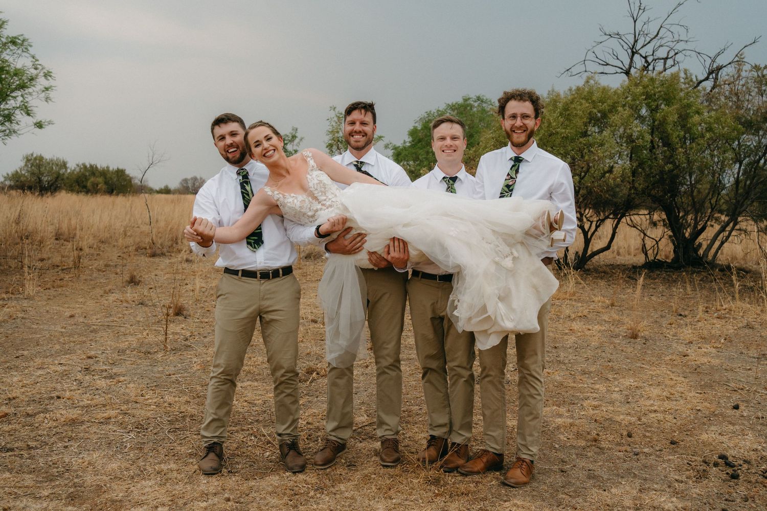 Wedding party in casual attire poses playfully in a rustic outdoor setting with winter dried grass and trees.