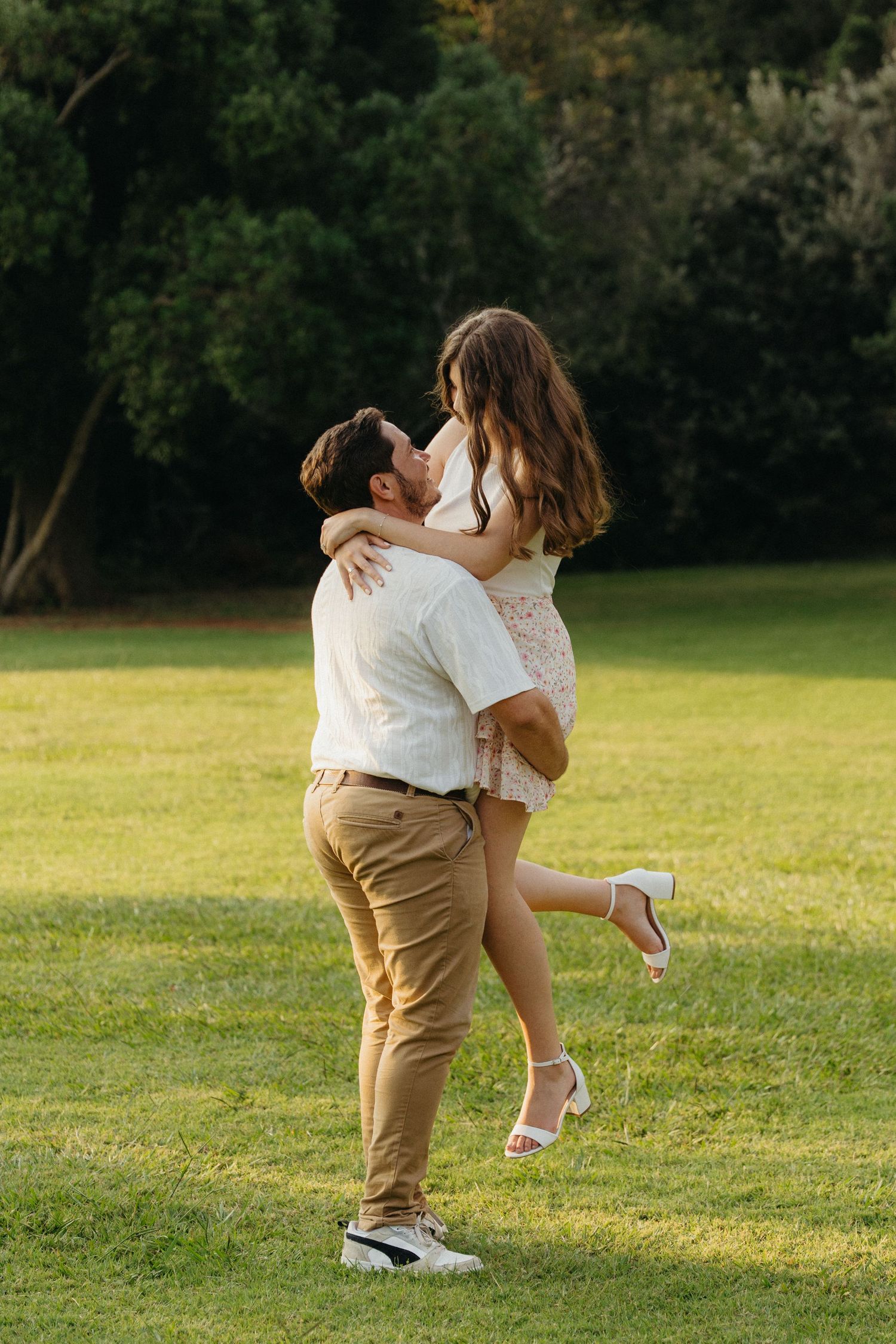Couple embracing on green lawn, man lifting woman in white dress, romantic outdoor moment in sunlit park.