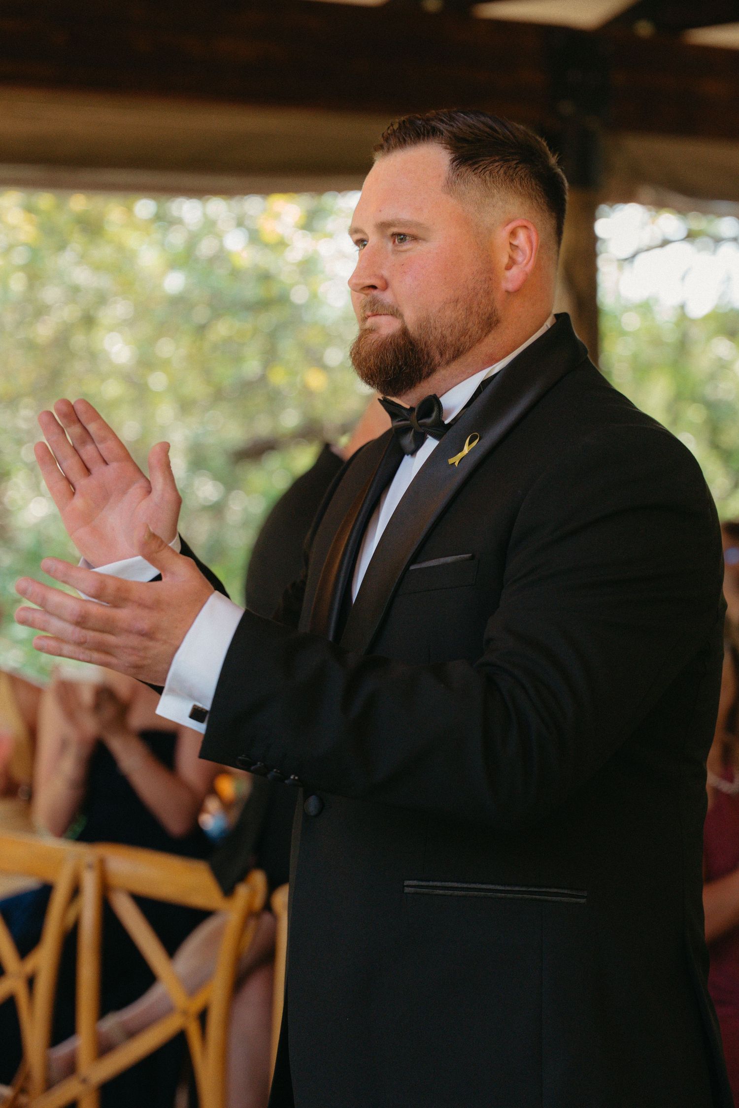 Men in black suits applaud during a formal indoor event with wooden chairs visible in background.