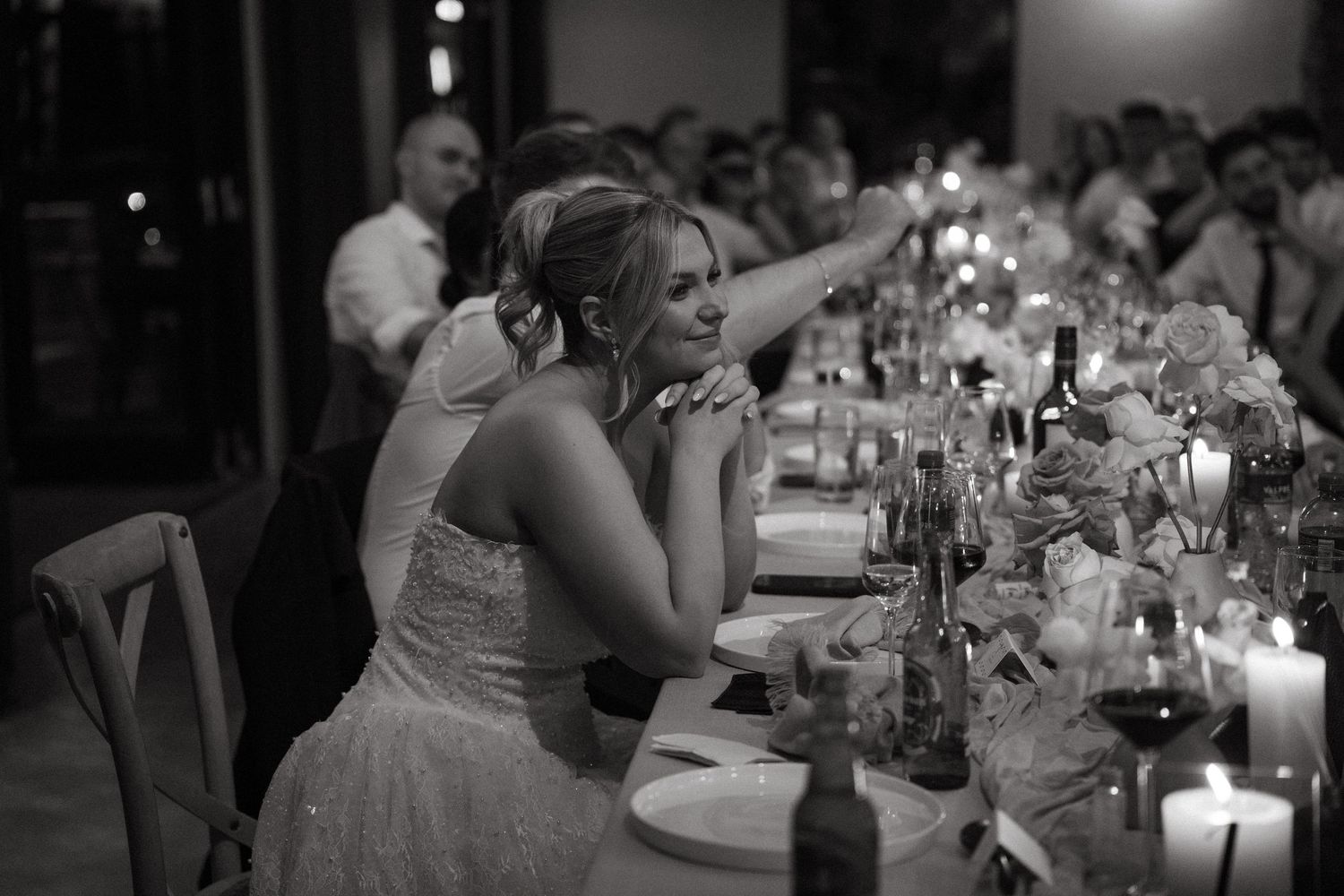 A black and white candlelit wedding reception shows someone in a white dress seated at an elegantly decorated dining table.