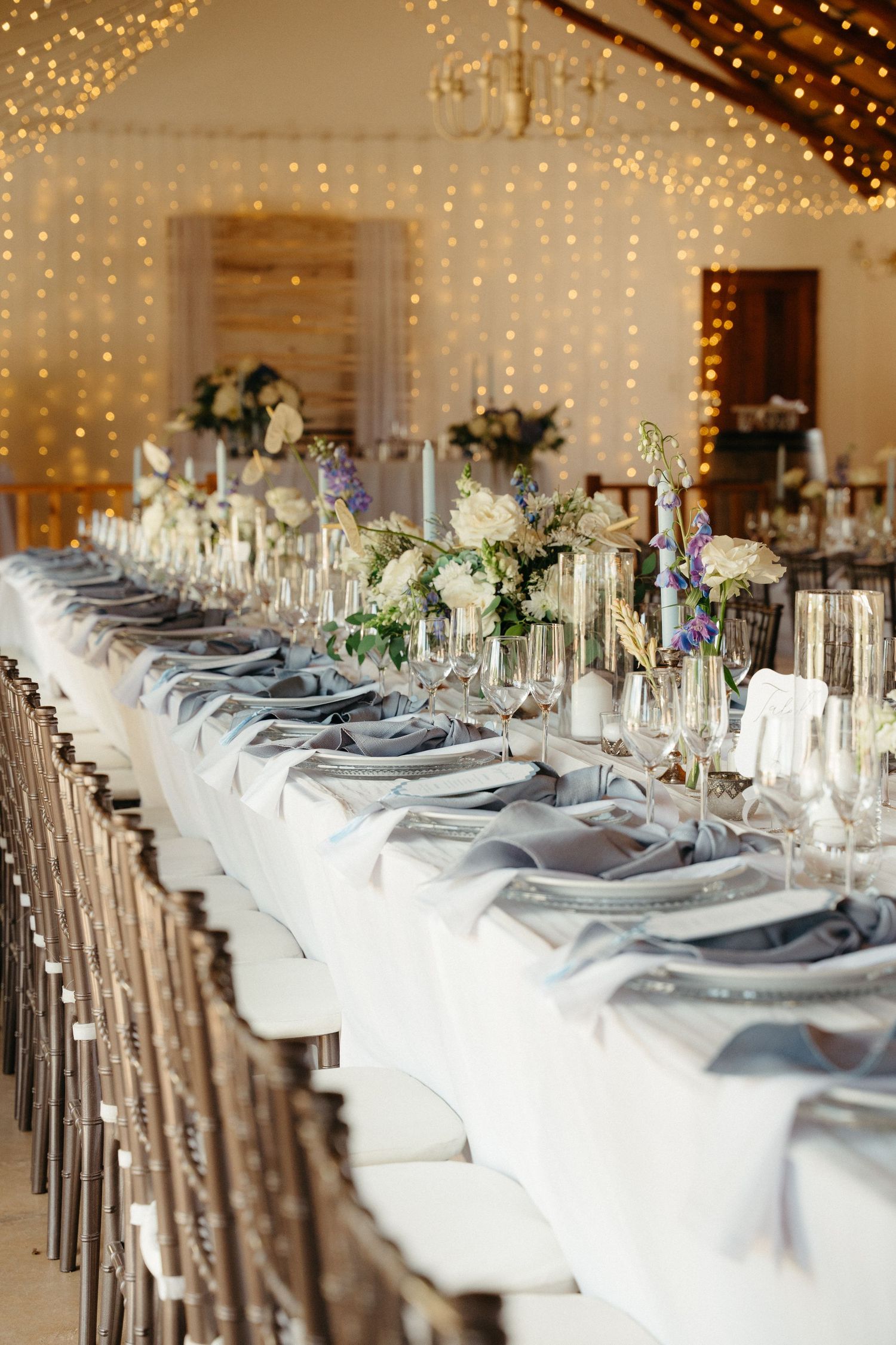 Elegant wedding reception tables set with white linens, gray napkins and rustic chairs under twinkling string lights.