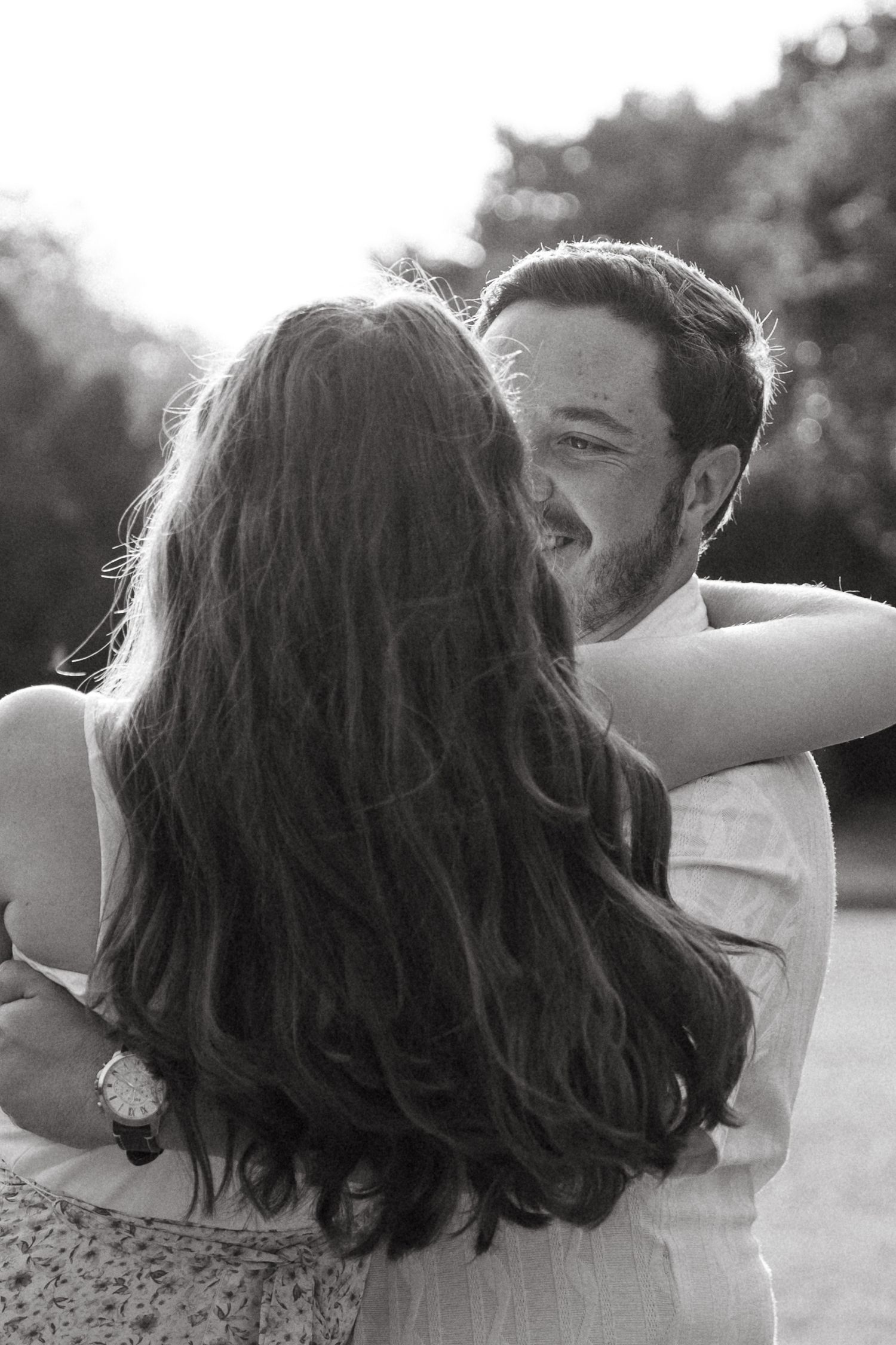 Black and white photo of a couple embracing outdoors, woman with long wavy hair, man smiling warmly in soft sunlight.