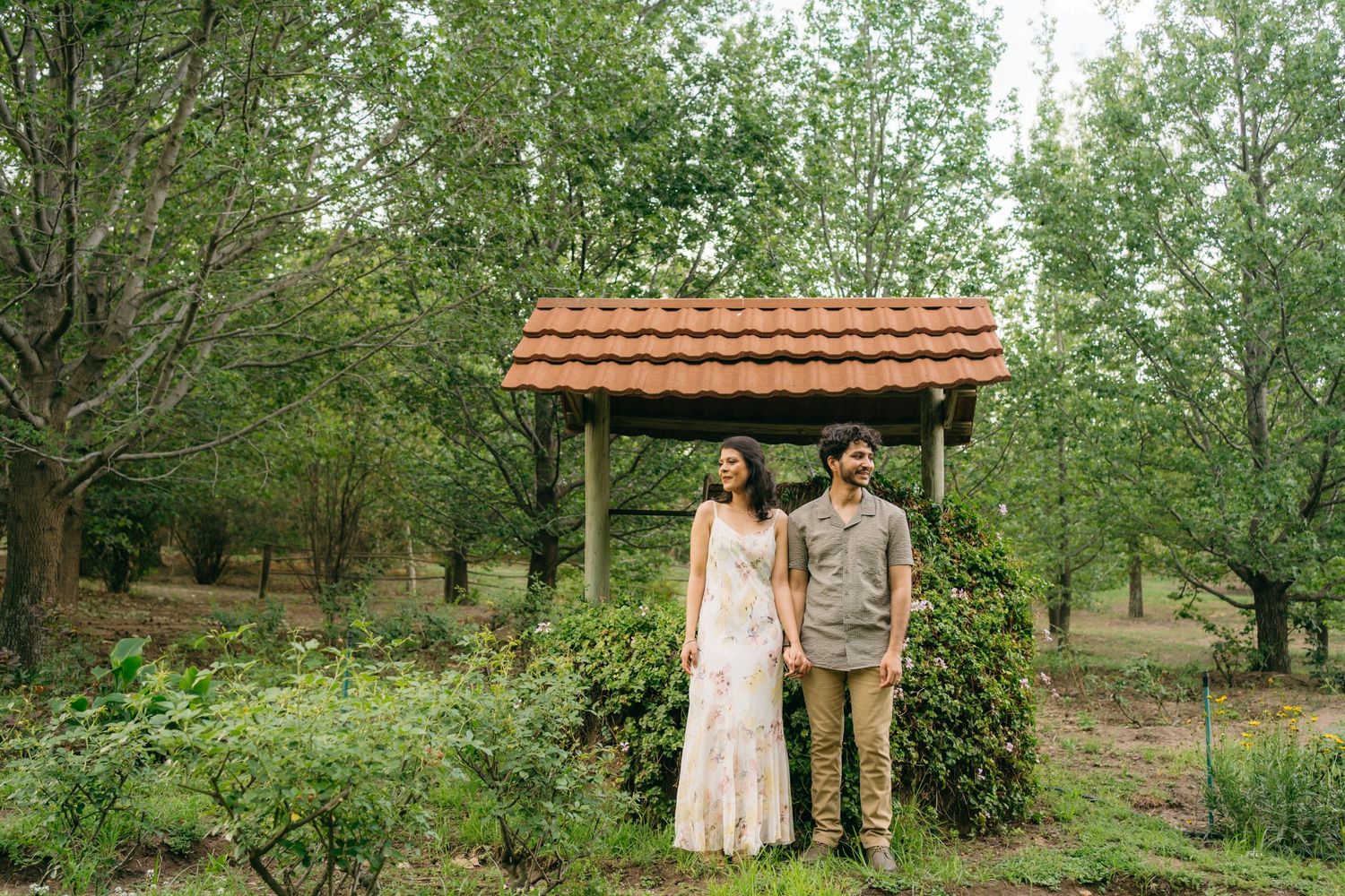A couple stands under a rustic wooden gazebo in a garden setting.