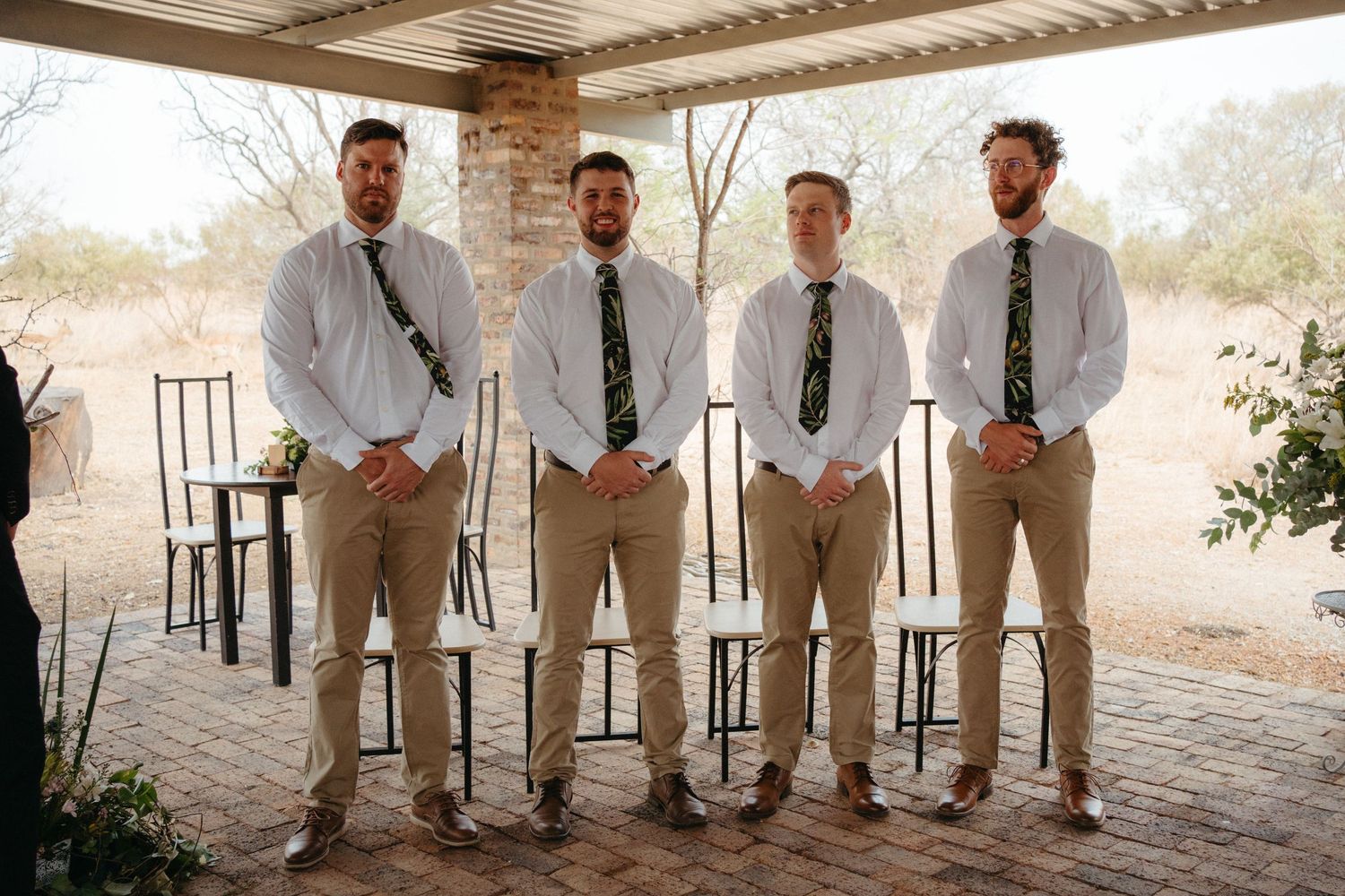 Four groomsmen in matching khaki pants and dress shirts pose on a covered brick patio.