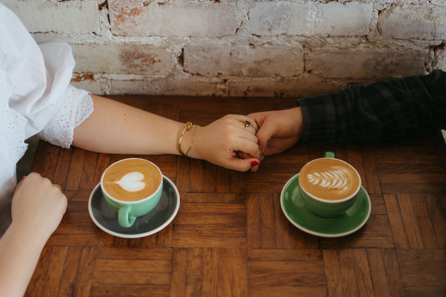 Two green ceramic cups of latte art on a wooden table with hands resting nearby.