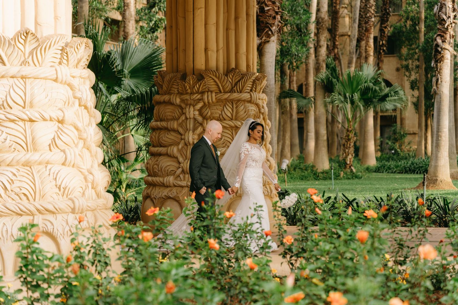 Bride and escort walk through lush garden with orange roses and tall palm trees at Sun City