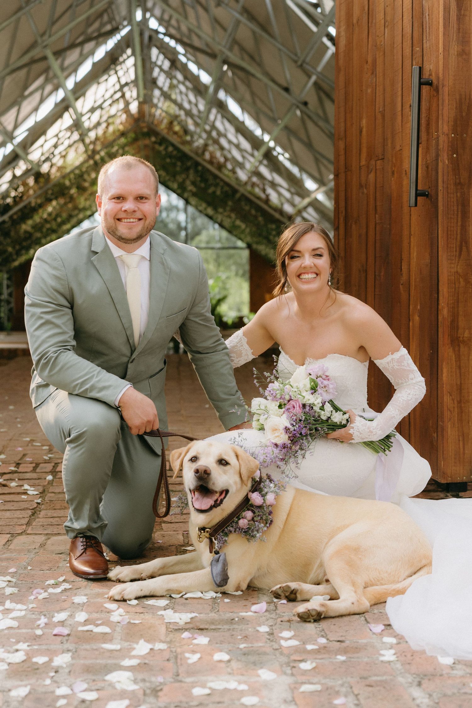 Happy newlyweds posing with their yellow Labrador dog in a rustic greenhouse venue.