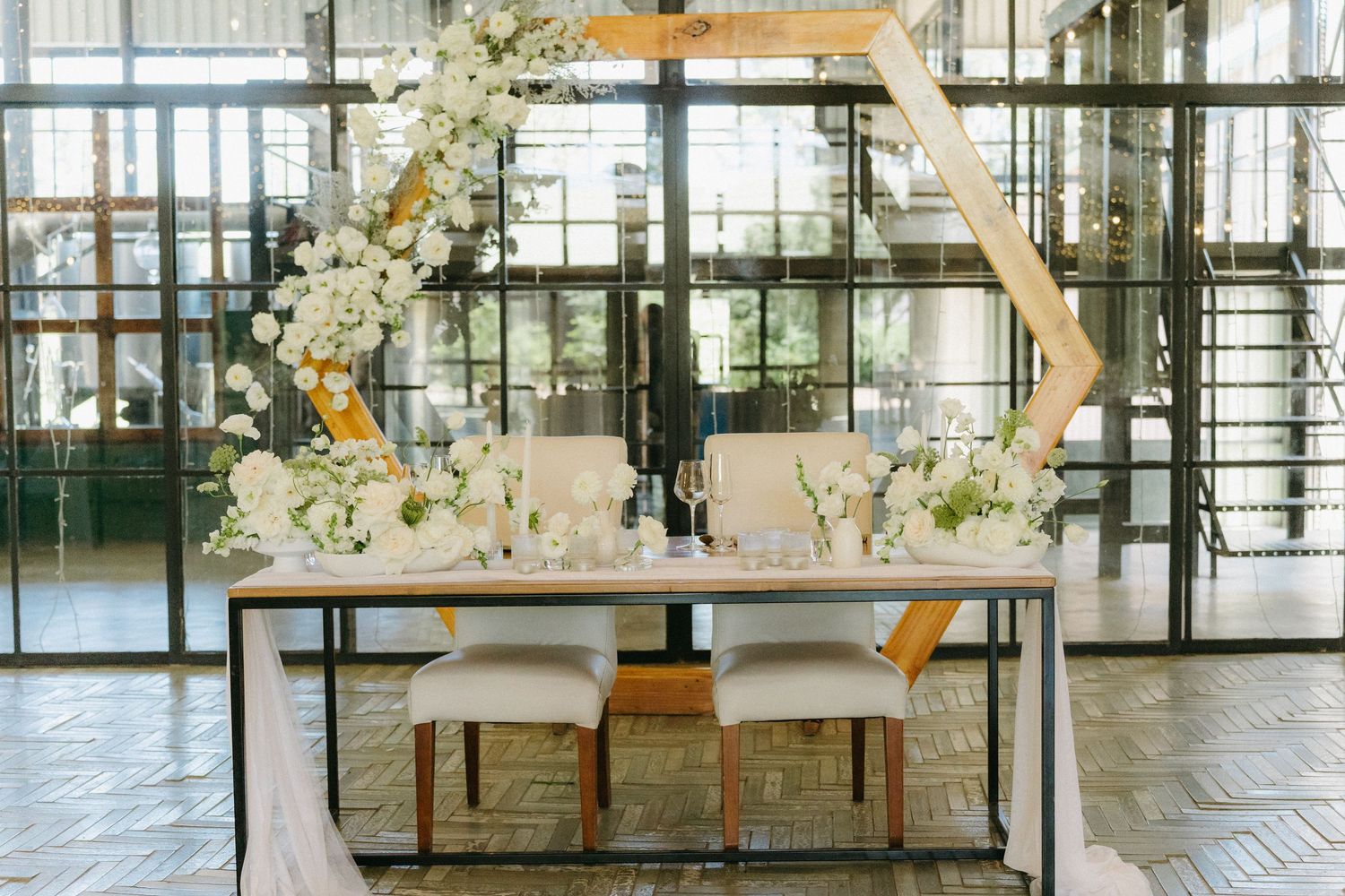 Modern wedding reception setup with hexagonal arch and white floral arrangements in an industrial-style venue.