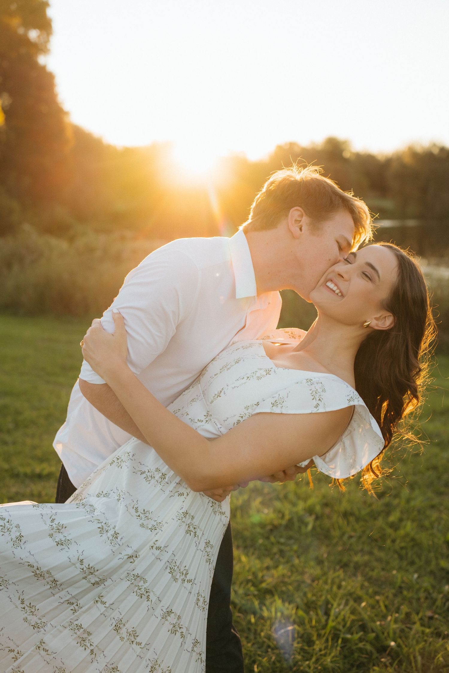 A couple shares a romantic embrace in a floral dress and white shirt at sunset in a grassy field with golden light.