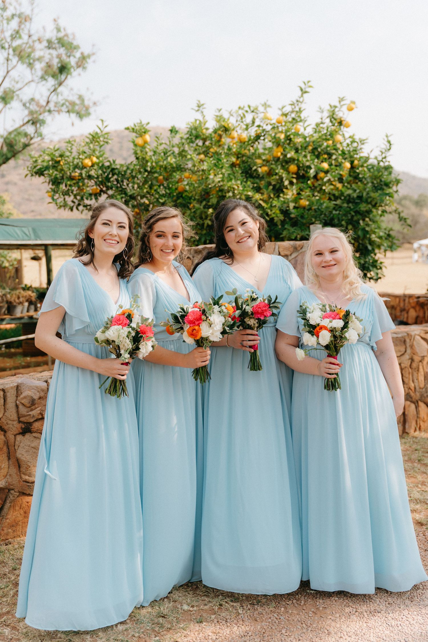 Bridesmaids in light blue floor-length dresses holding colorful bouquets pose together against a desert landscape.