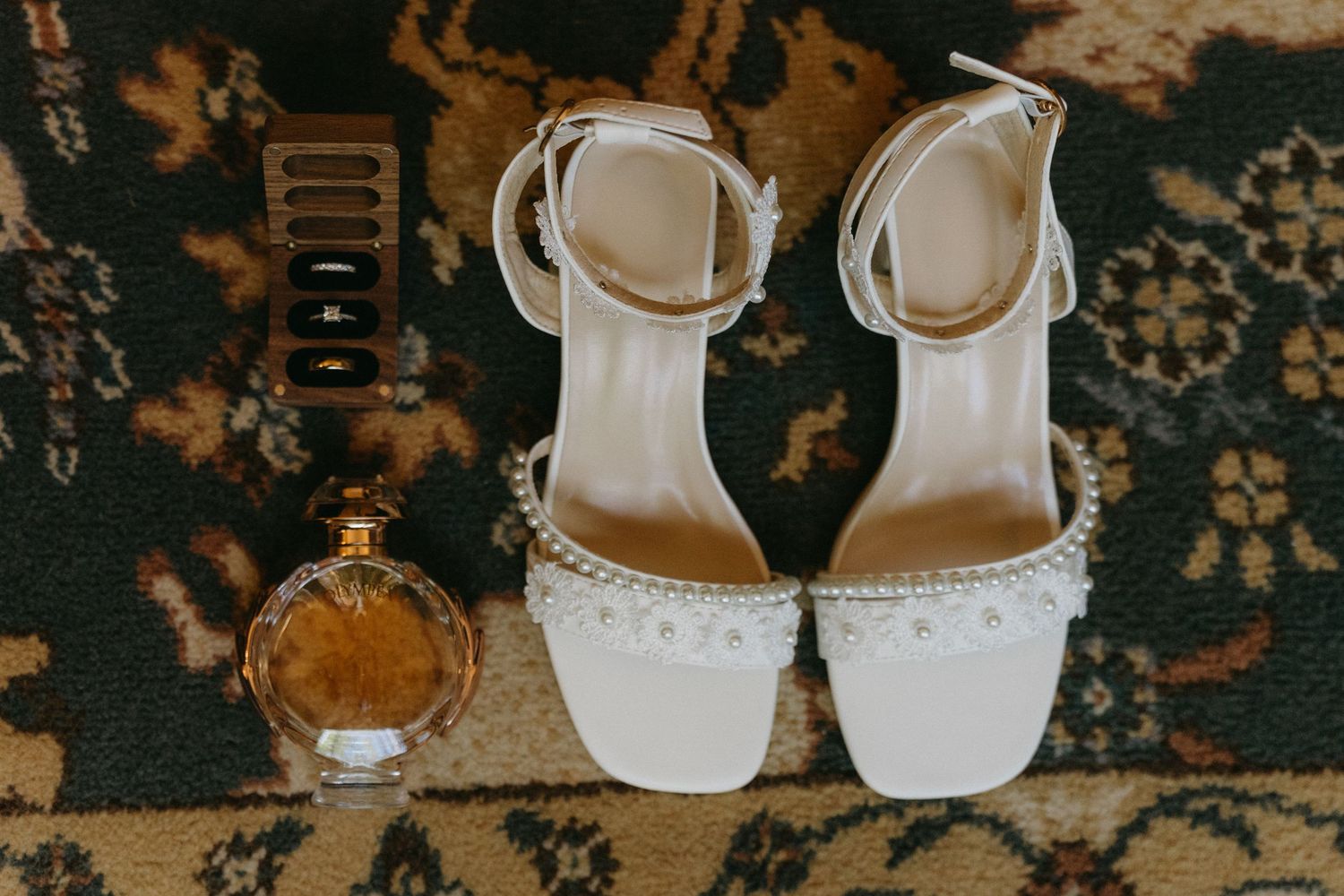 White wedding sandals with ankle straps and pearls sit next to a round perfume bottle on a patterned dark carpet.