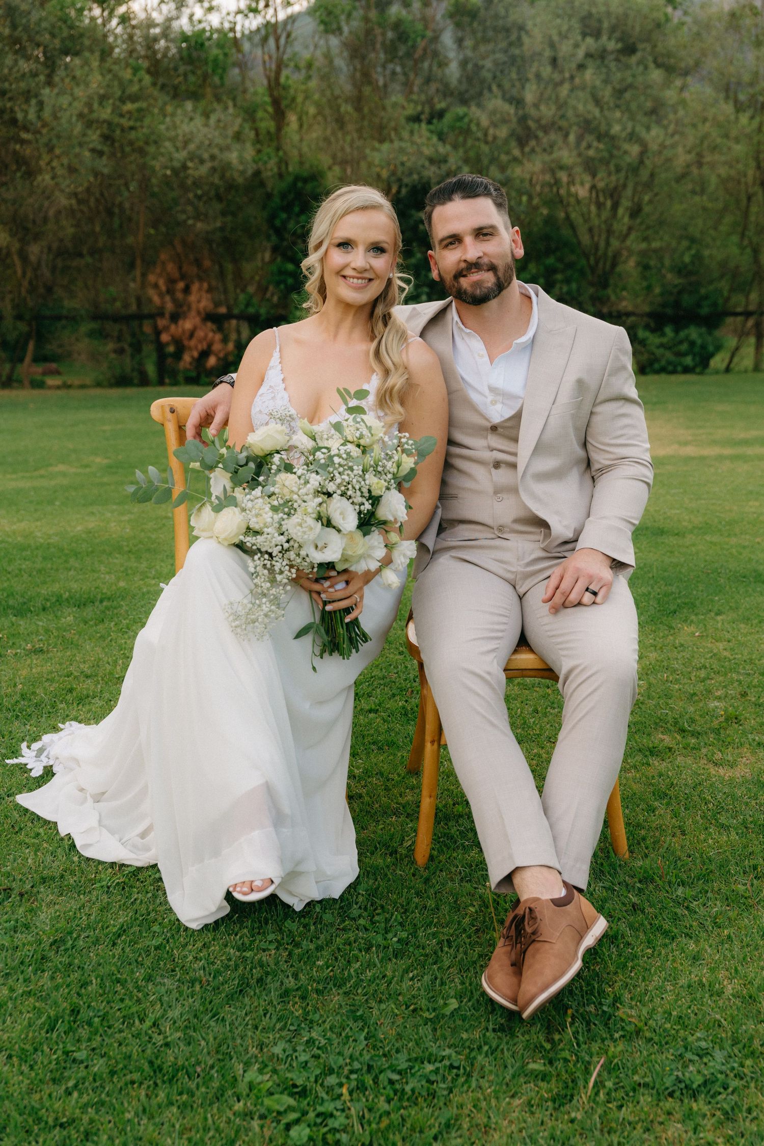 Wedding couple sits together on wooden chairs in a garden setting with white floral bouquet.