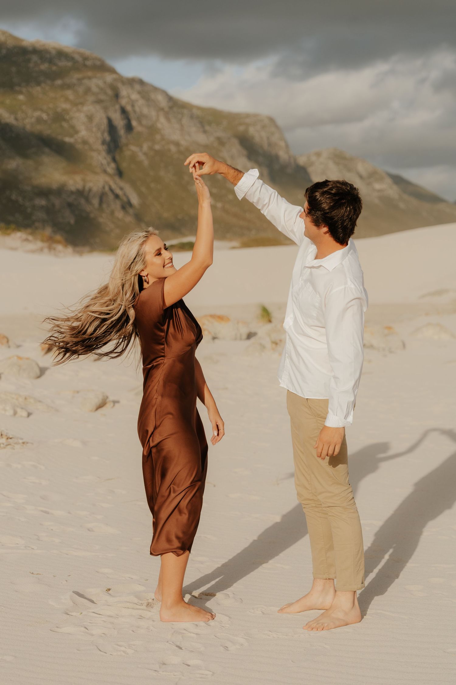 Couple dancing barefoot on white sand dunes with mountains in background, woman in brown silk dress, man in white shirt.