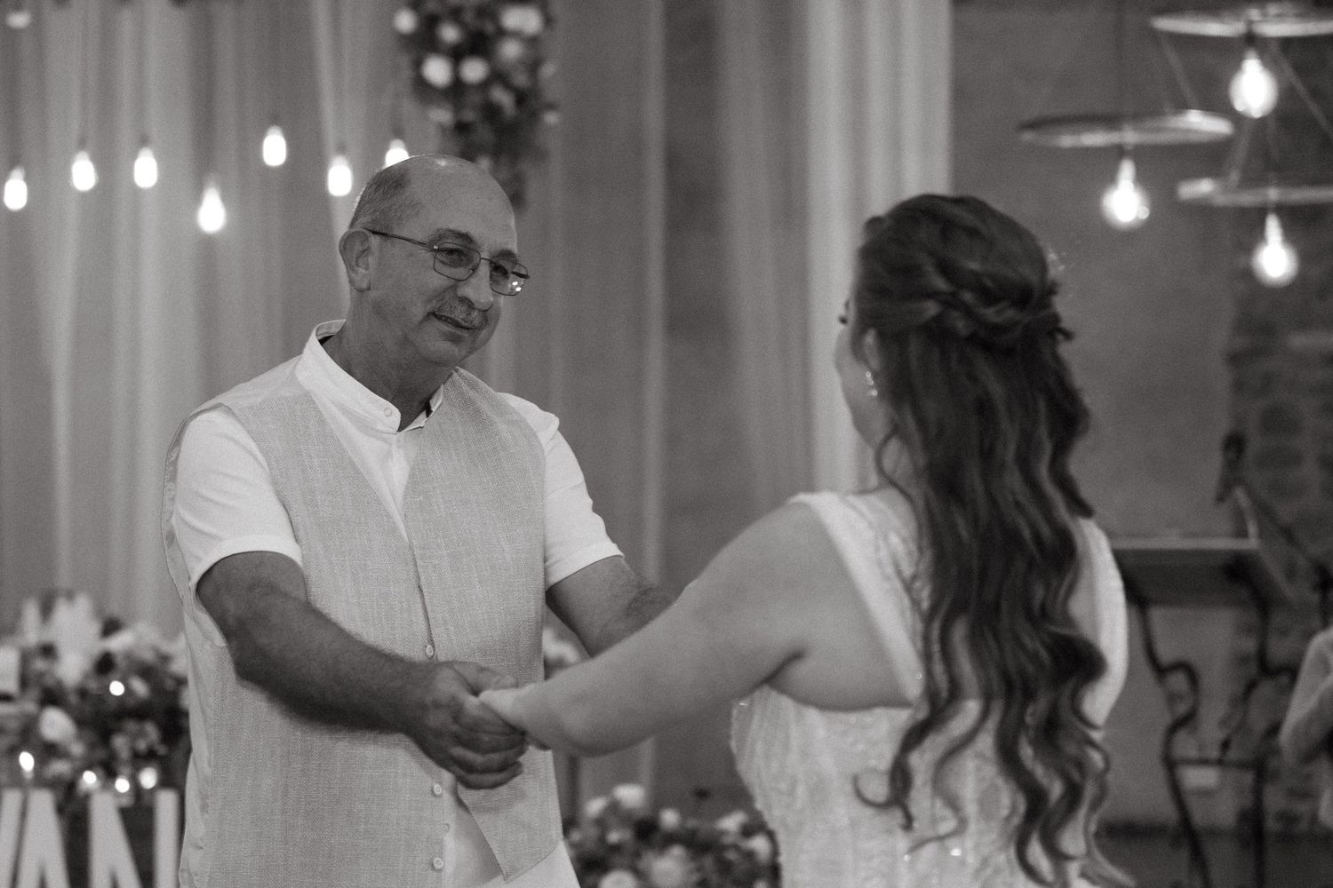 Black and white photo of couple dancing during wedding reception with hanging lights.