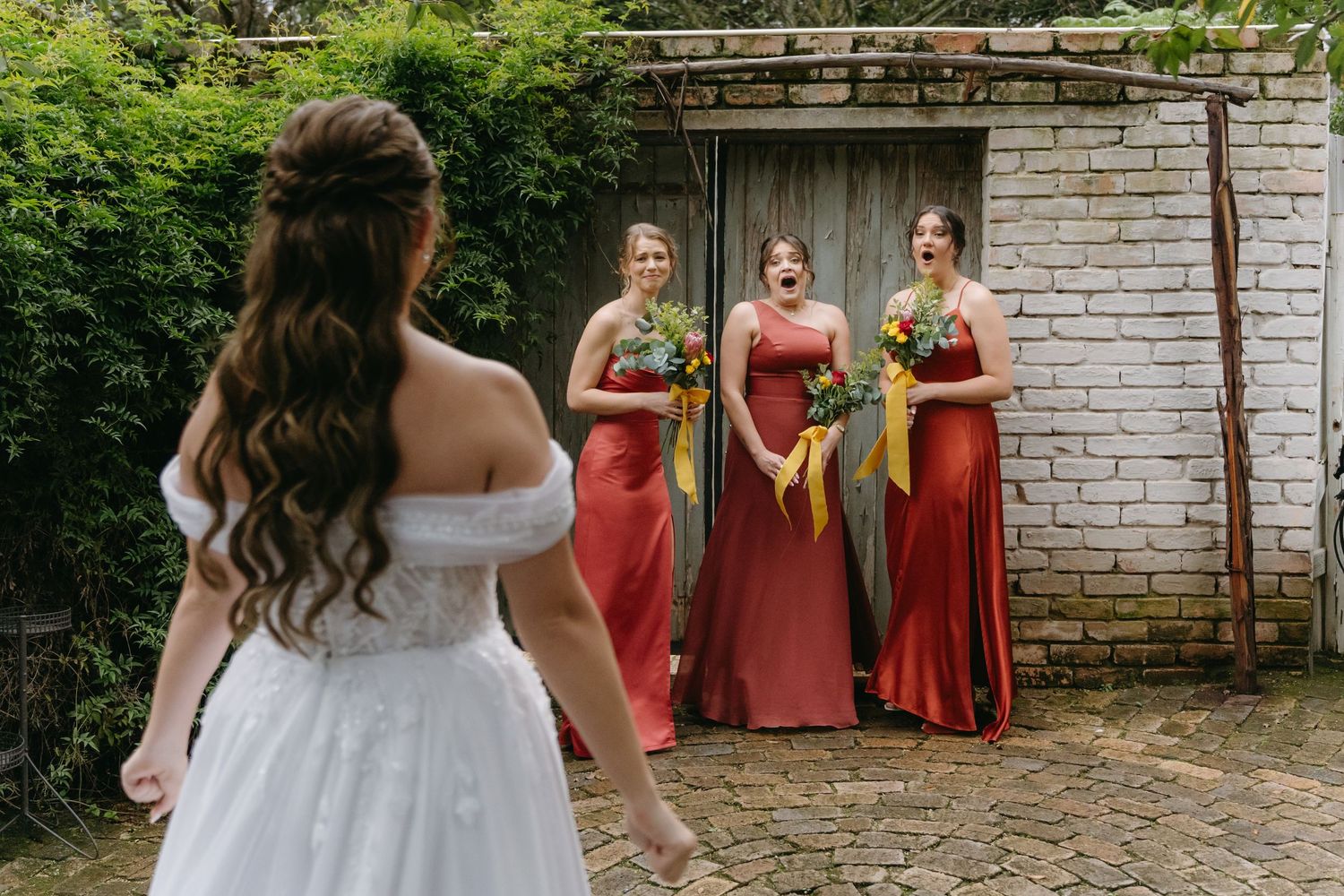 Back view of bride in white dress facing bridesmaids in coral gowns against brick wall.