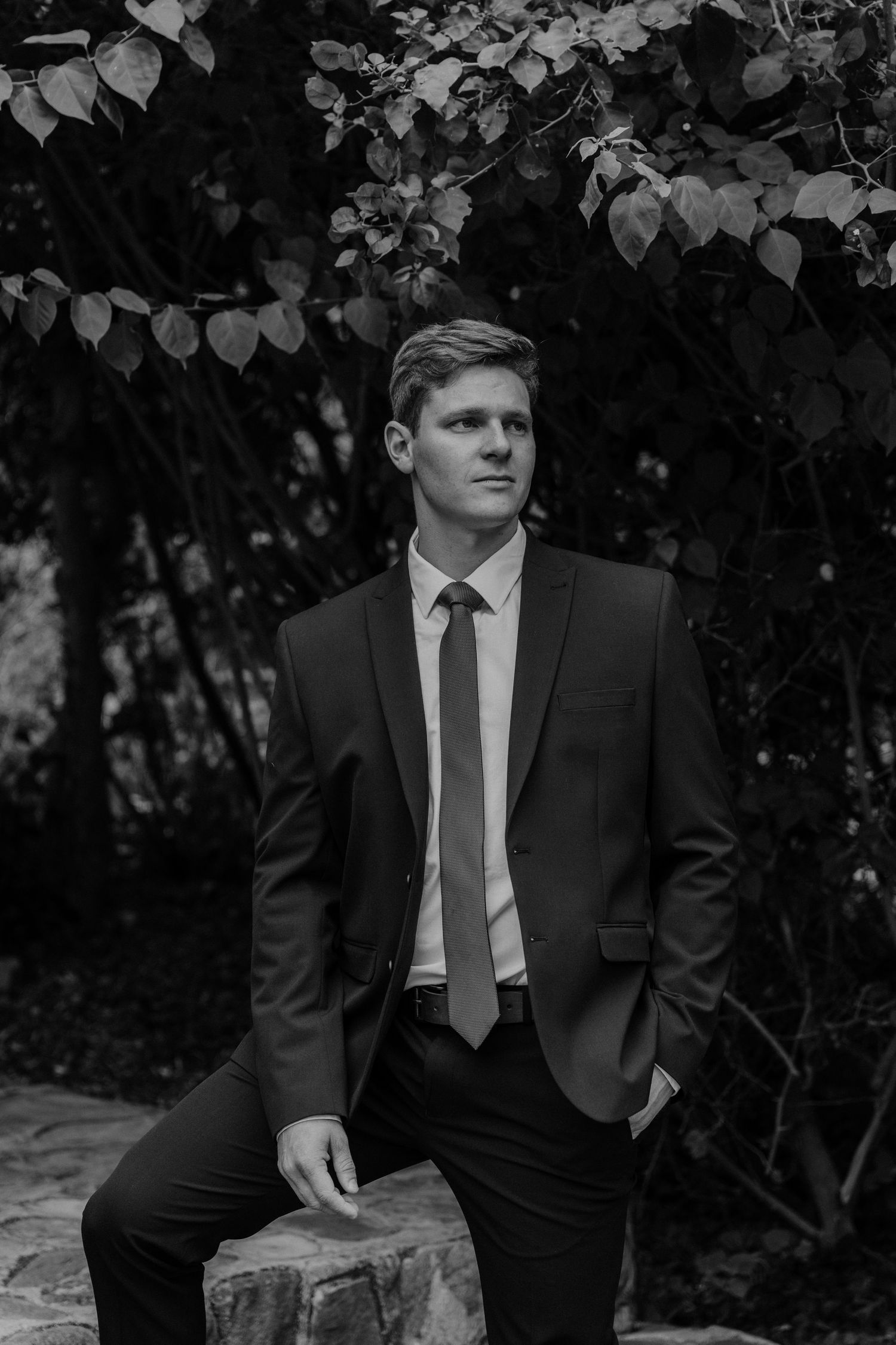 A person in a black suit and tie poses in a garden setting with dramatic black and white lighting.