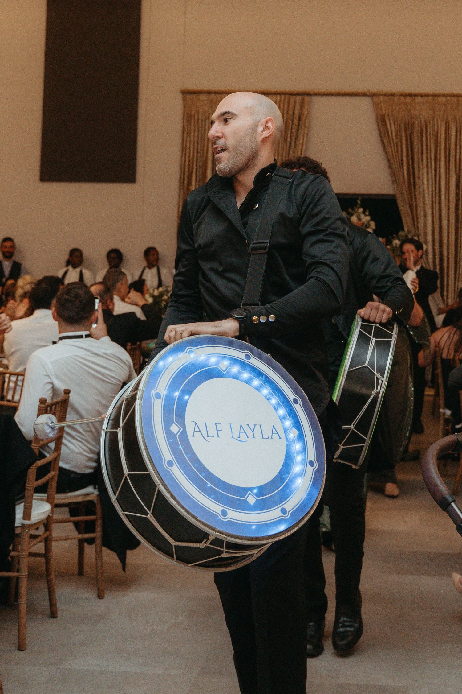 Bald male drummer performing with illuminated Alf Layla drum at elegant indoor wedding reception event.