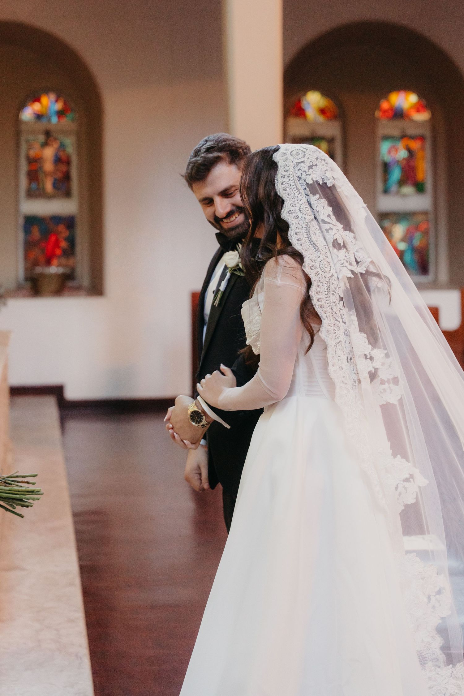 Bride and groom share a joyful moment at the church altar, with stained glass windows in the background.