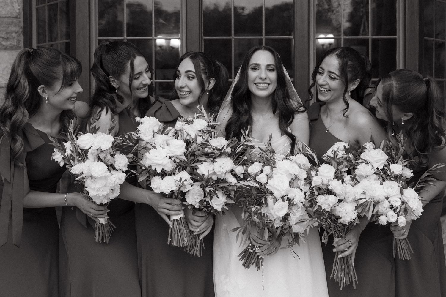 Black and white photo of a bride and five bridesmaids laughing together while holding white floral bouquets.