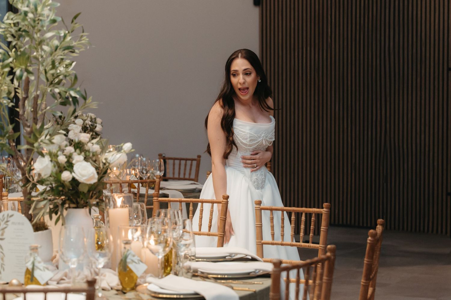 Bride in white gown stands at elegantly decorated wedding reception table with candles and floral centerpieces.