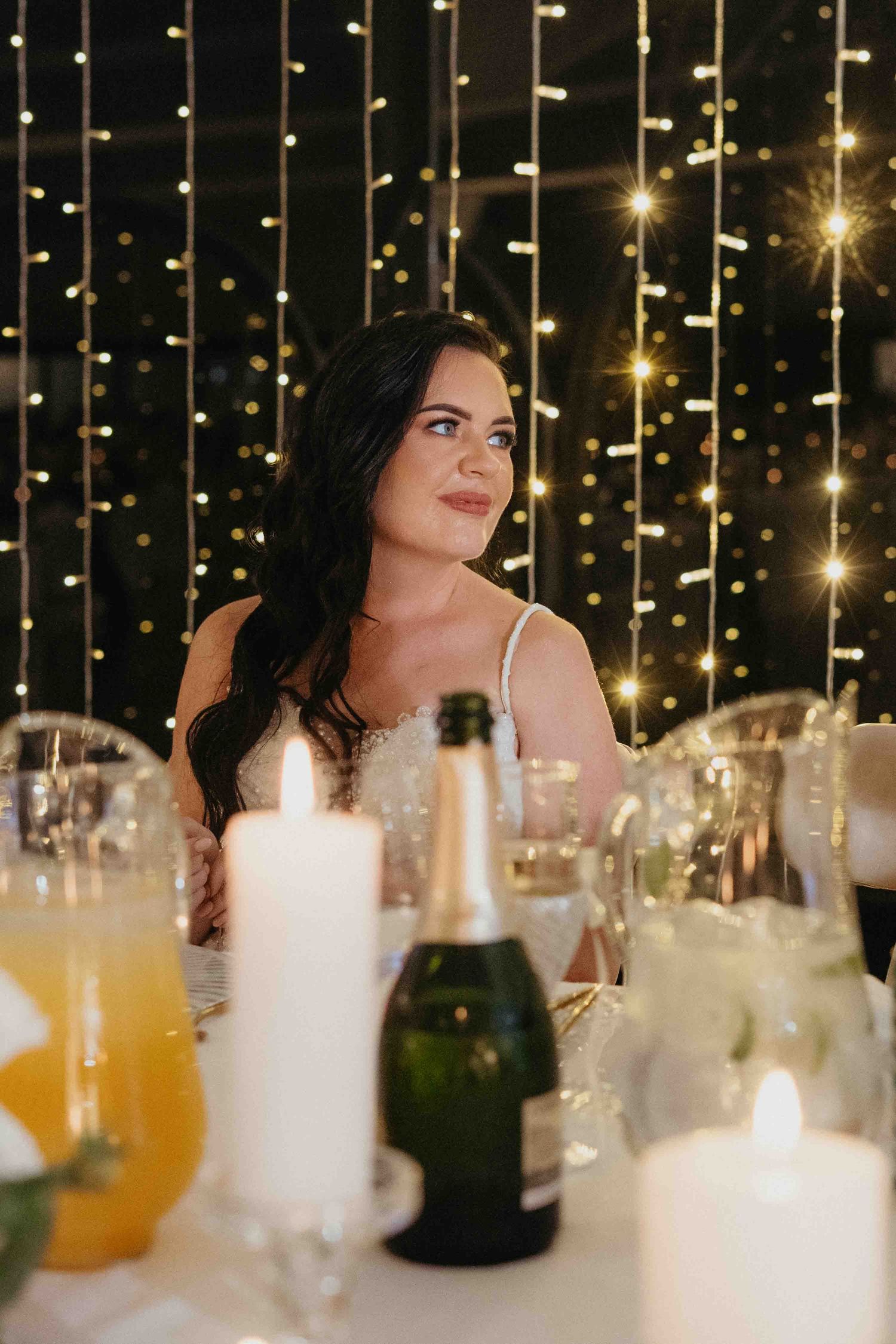 A bride sits at a candlelit reception table with champagne, smiling softly against a backdrop of twinkling fairy lights.