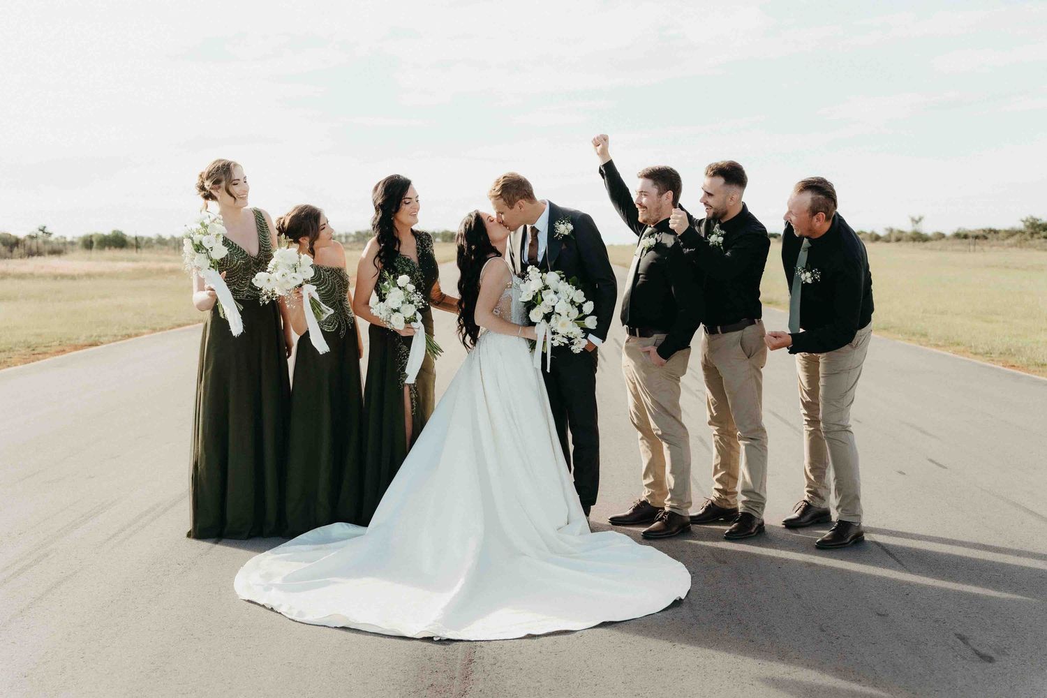 Bride and groom kiss surrounded by bridesmaids in green dresses and groomsmen on an outdoor runway.