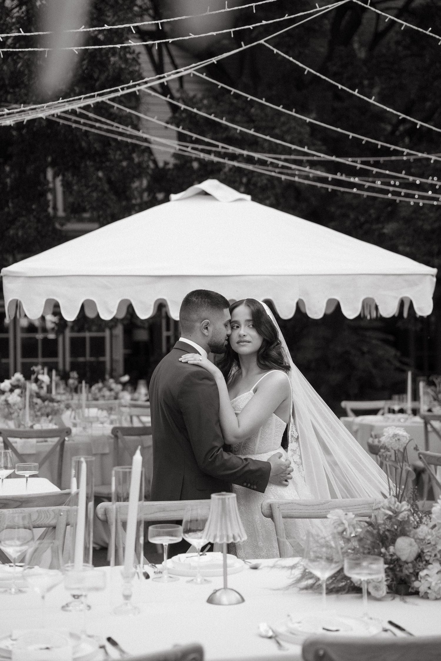 Black and white photo of bride and groom embracing outdoors under string lights near a white tent at a wedding reception.