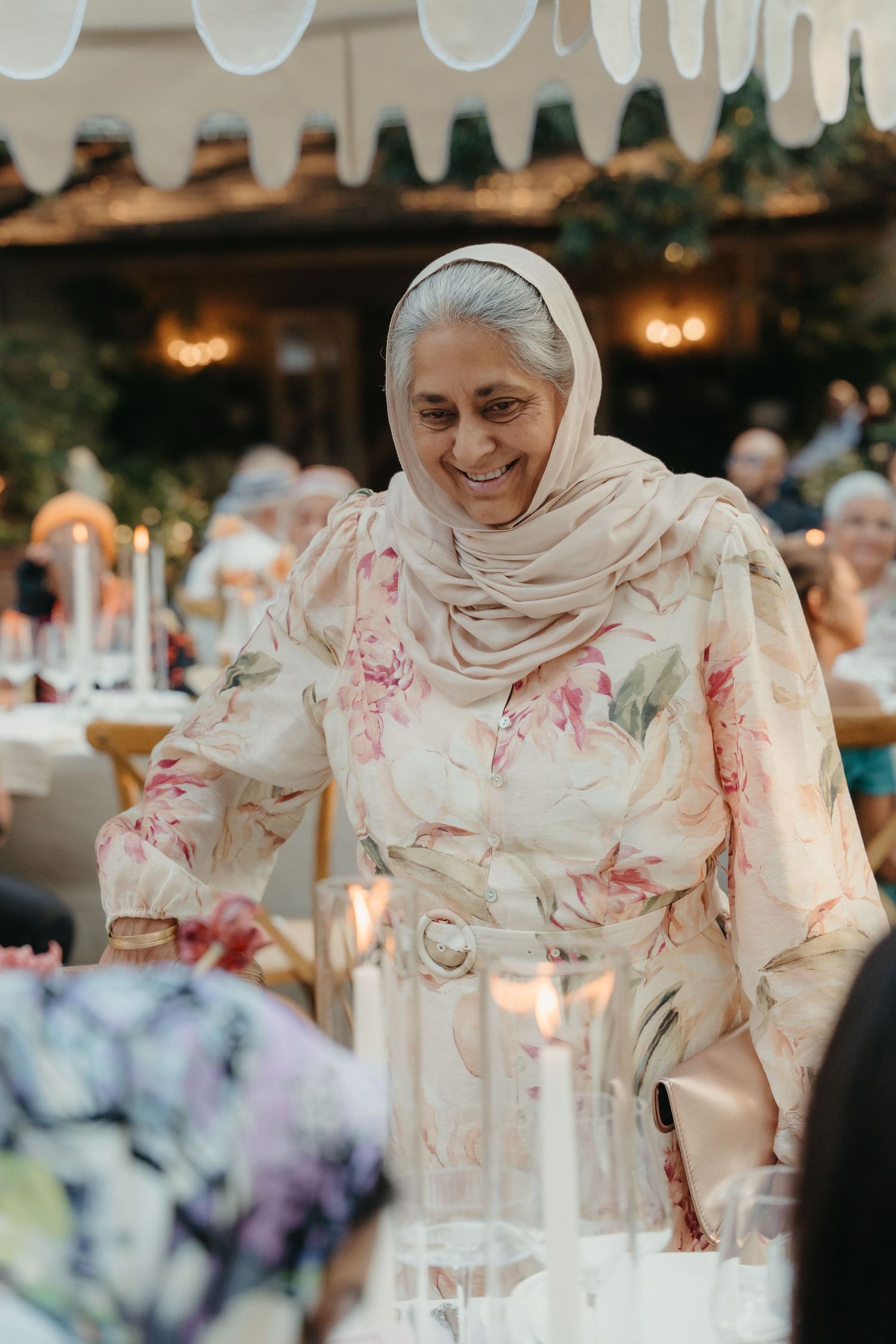 Smiling woman in floral dress and hijab at elegant wedding reception with candles and guests.