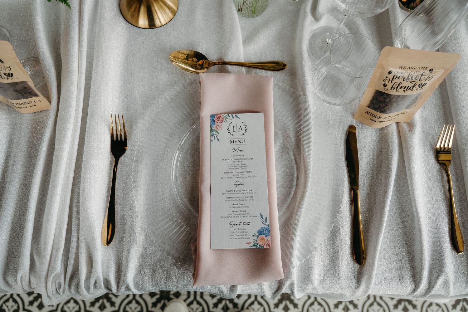 Elegant wedding table setting with gold cutlery, pink napkin, floral menu card, and white tablecloth.