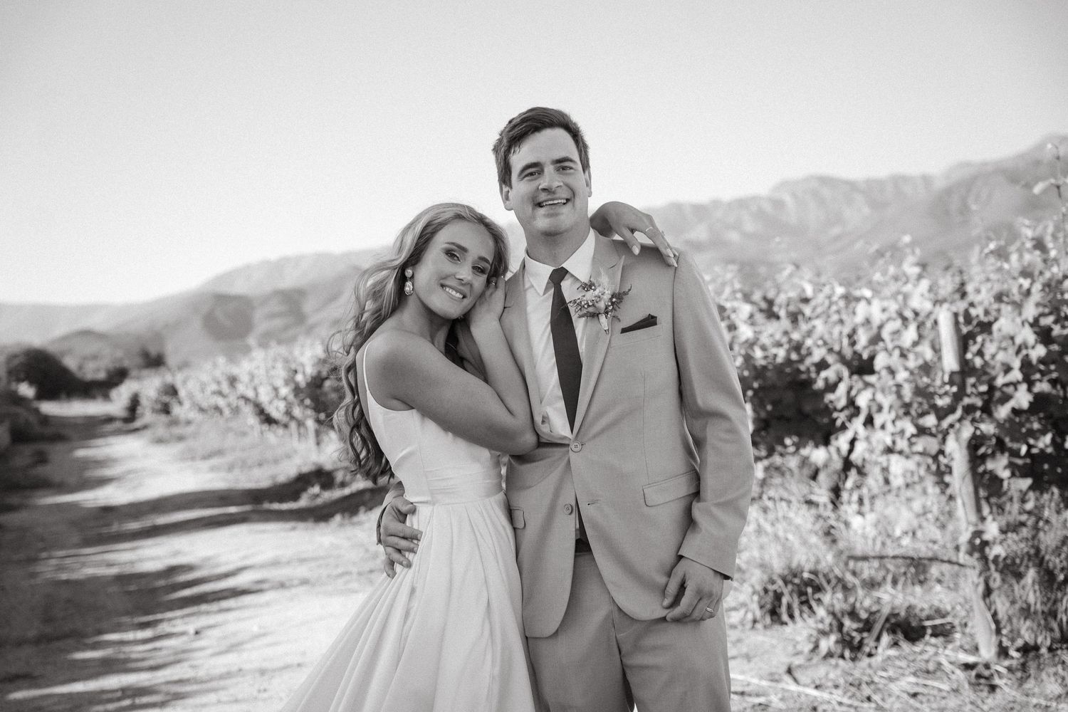 Smiling bride in a white gown embracing a groom in a light suit among Cape Town vineyard rows with mountains behind, in black and white.