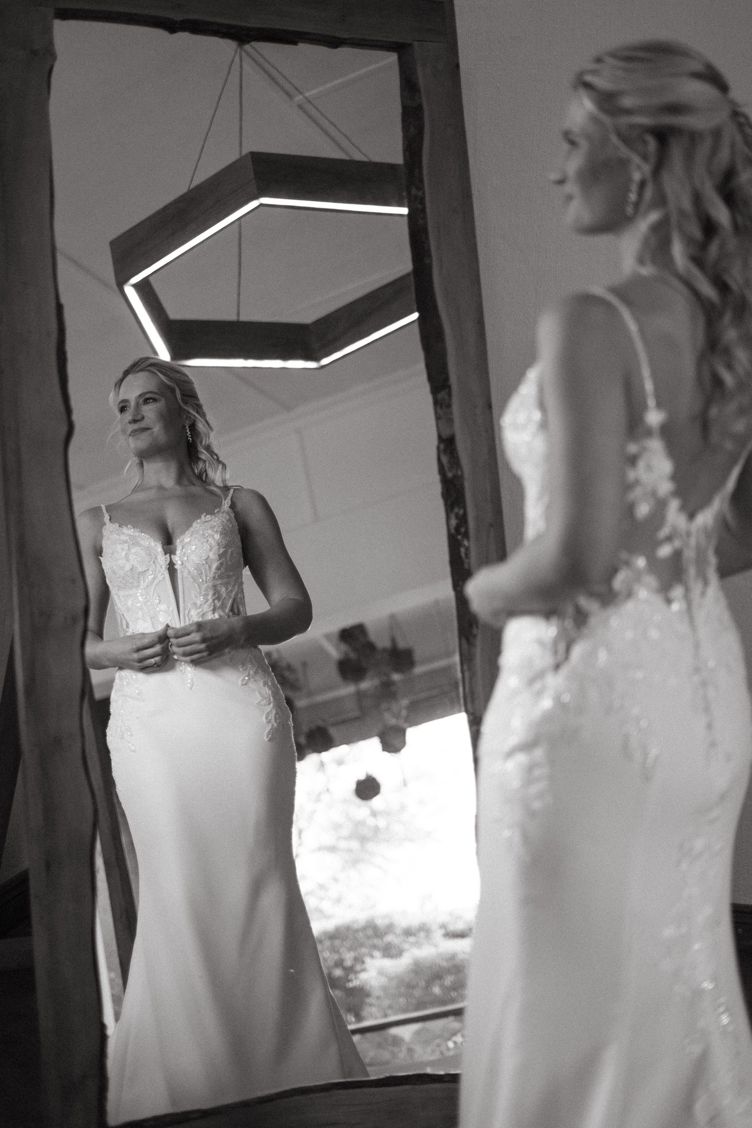 Bride in lace wedding gown admires her reflection in a large mirror, black and white photo.