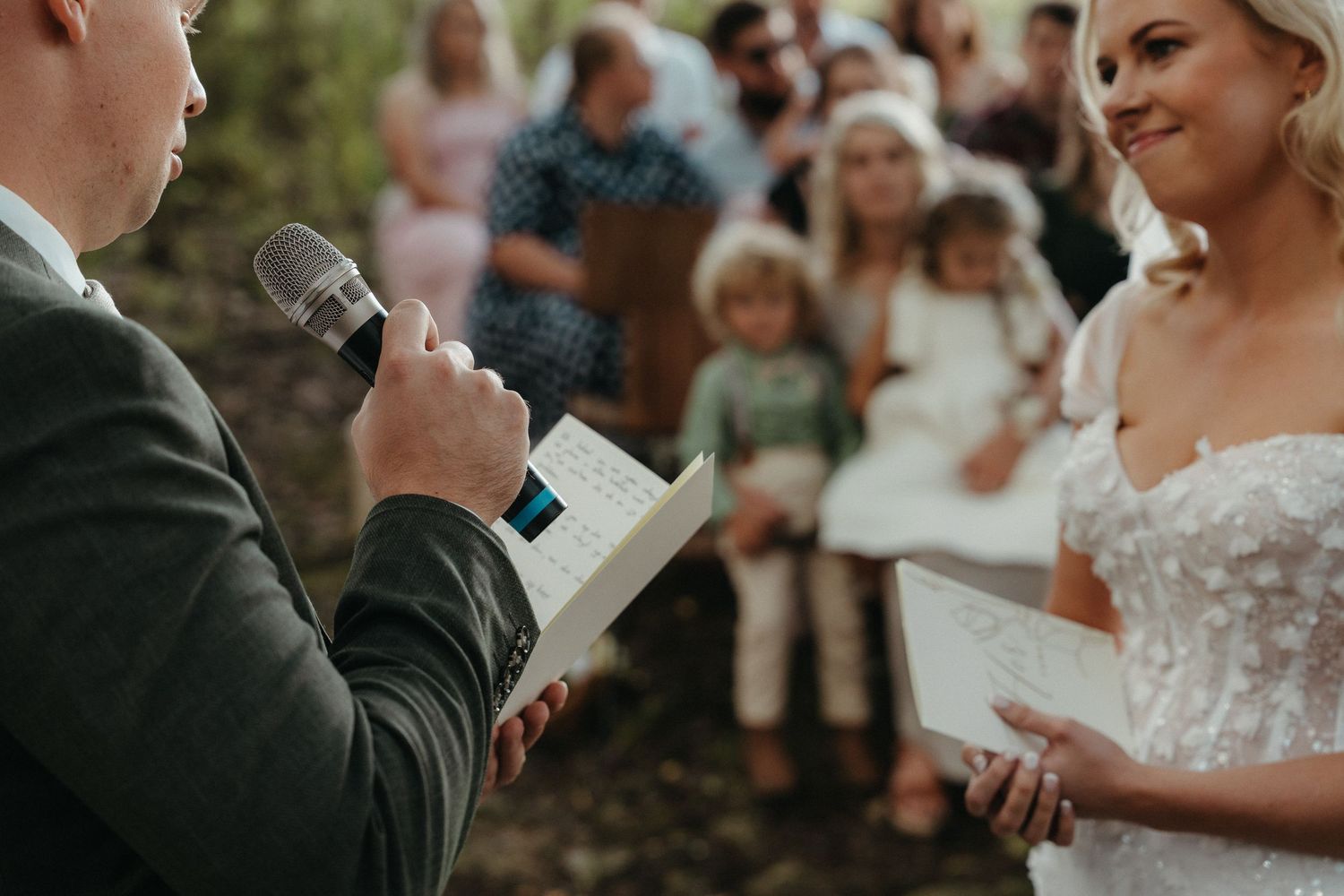 Groom reads wedding vows from a book at an outdoor ceremony while bride smiles in a lace dress.